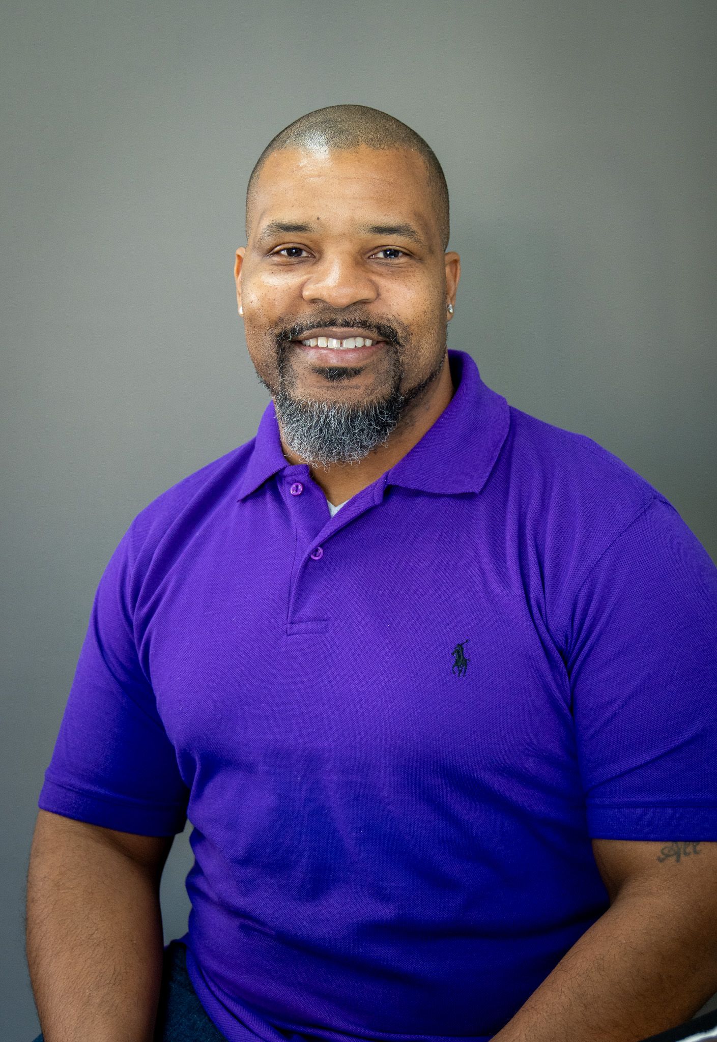 Professional headshot of person in purple polo shirt with gray beard against gray backdrop.