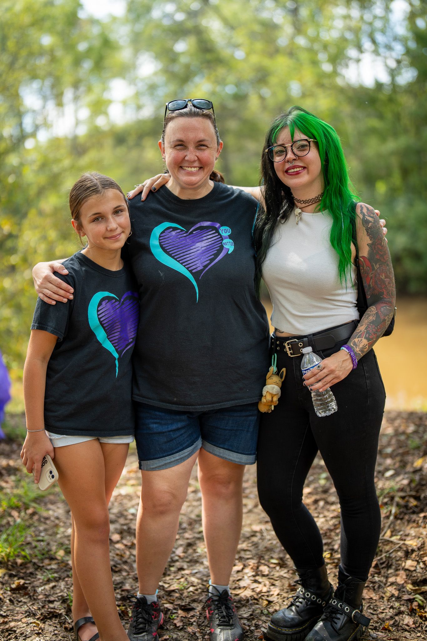 Three people pose together outdoors wearing matching purple heart t-shirts amongst trees in a casual summer setting.
