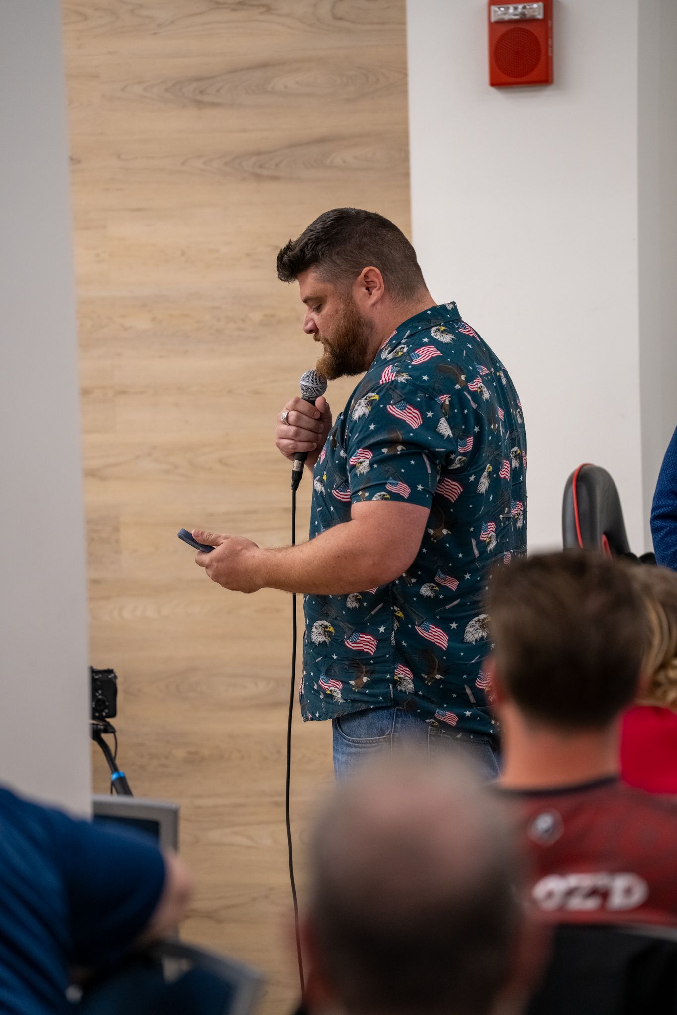 A person in a floral Hawaiian shirt stands speaking into a microphone while looking at their phone during a presentation.
