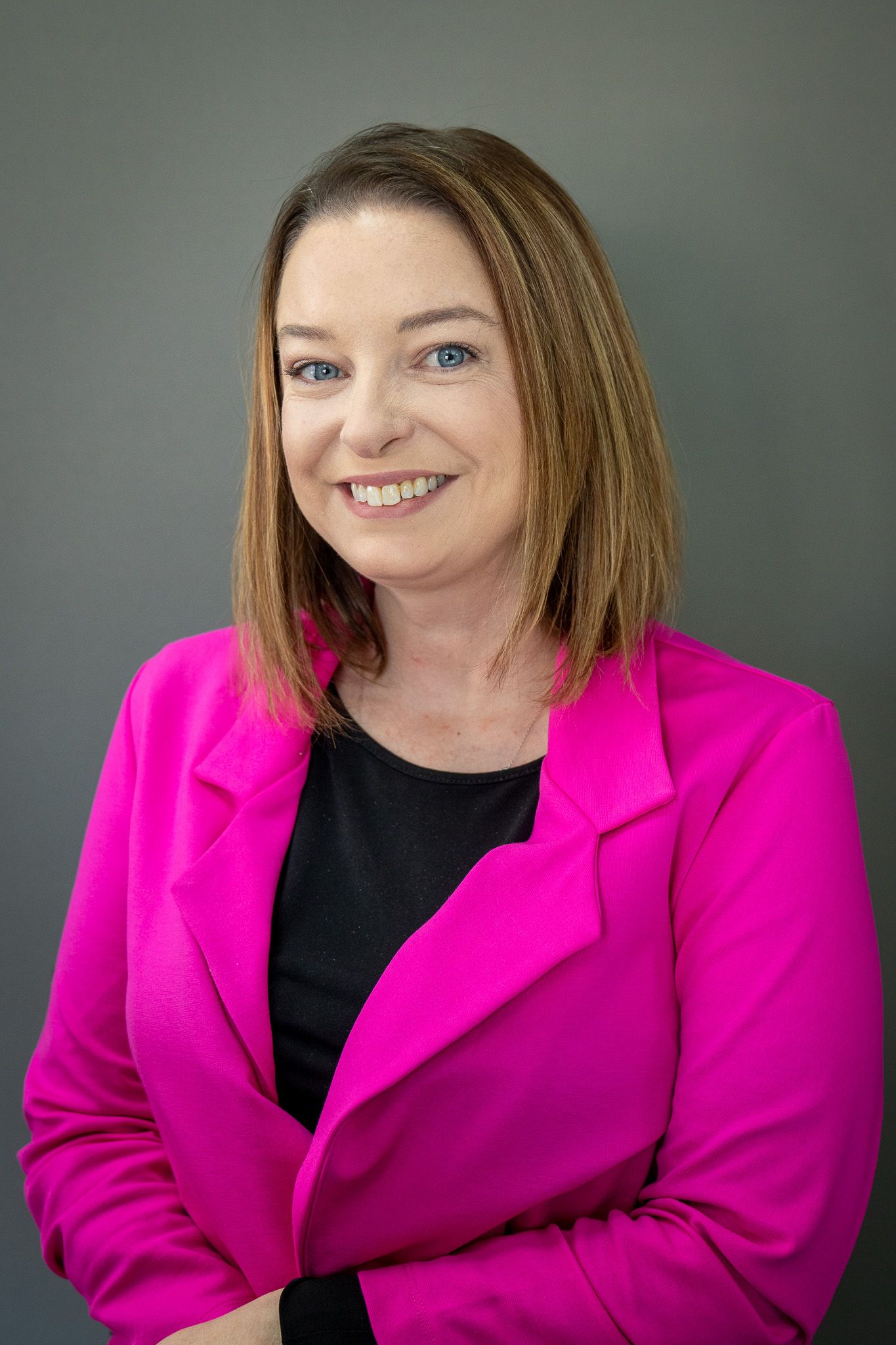 Professional portrait of person in bright pink blazer and black top smiling against grey backdrop.