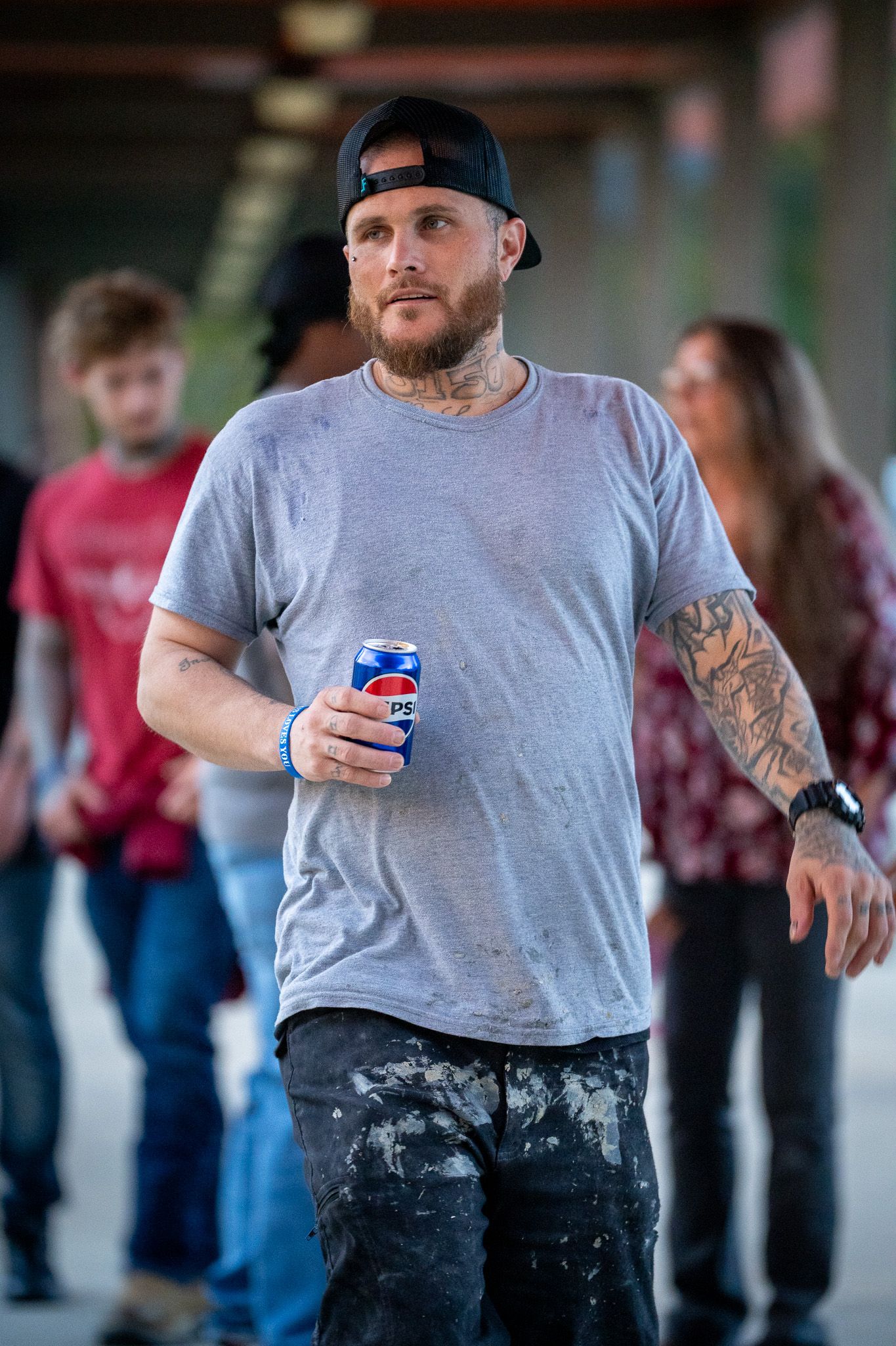 Person in a gray t-shirt and baseball cap holding a Pepsi can with tattoos on their arm while others stand in background.