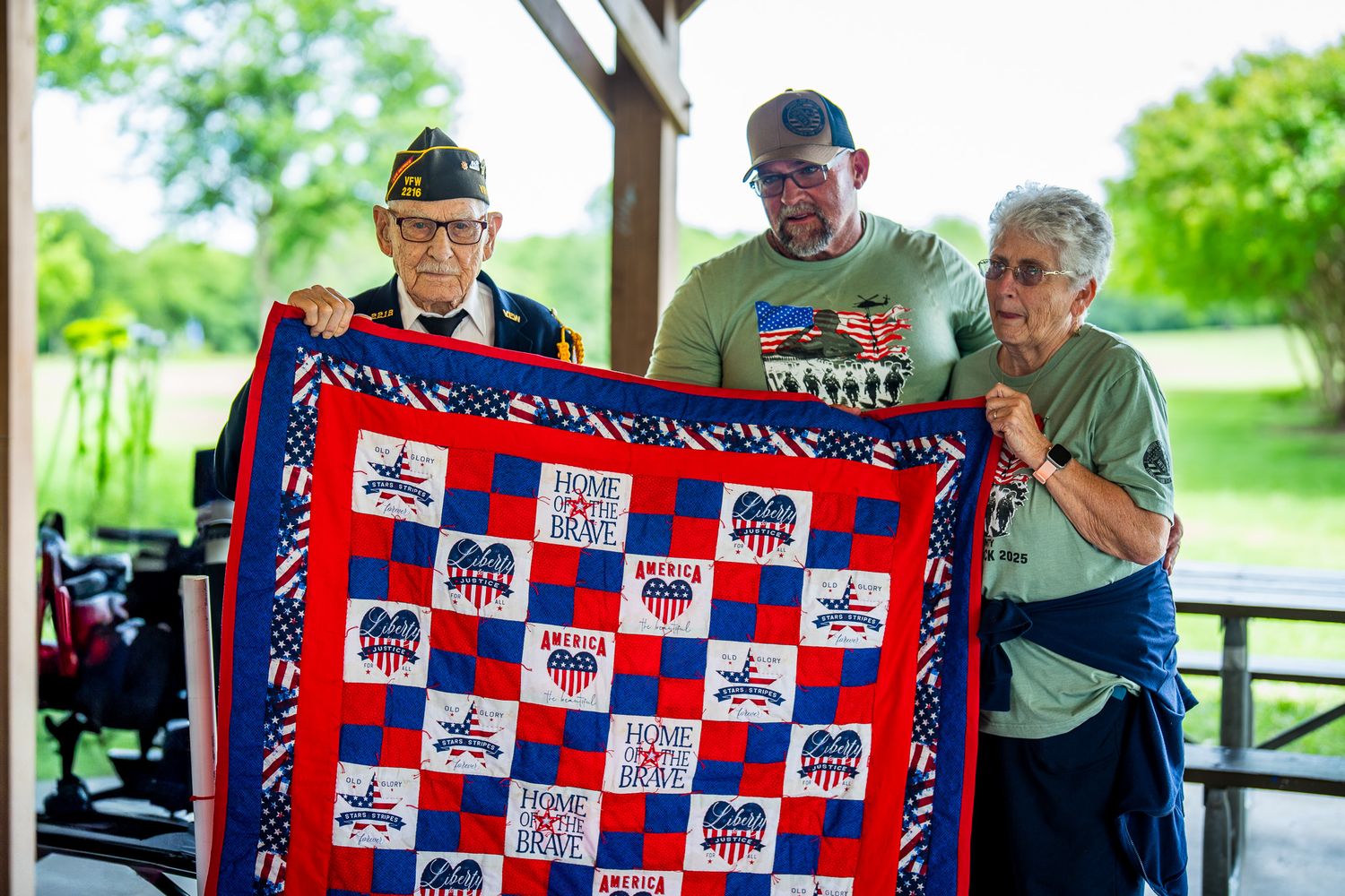 Three people displaying a patriotic red white and blue quilt with stars and stripes pattern at an outdoor gathering.