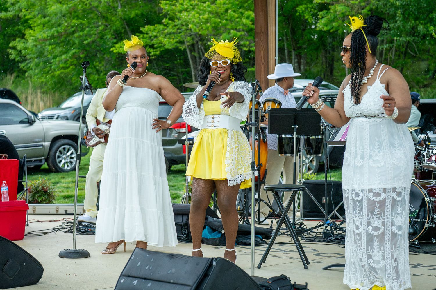 Musicians perform at an outdoor summer event wearing white and yellow dresses with decorative star accessories on stage.