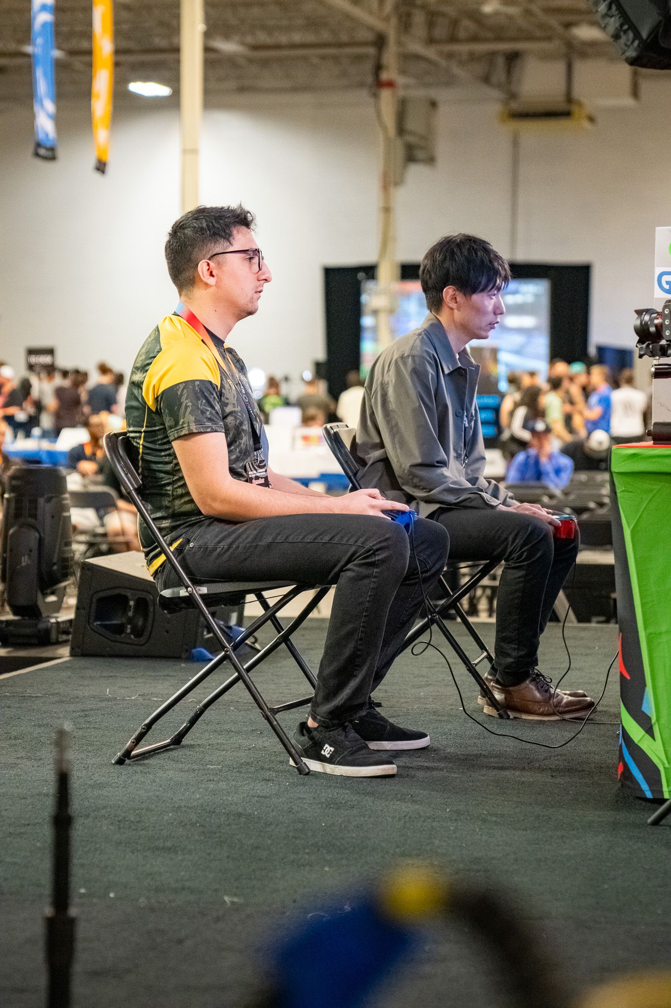 Two gamers seated on folding chairs focus intently during a competitive esports tournament in an indoor venue.