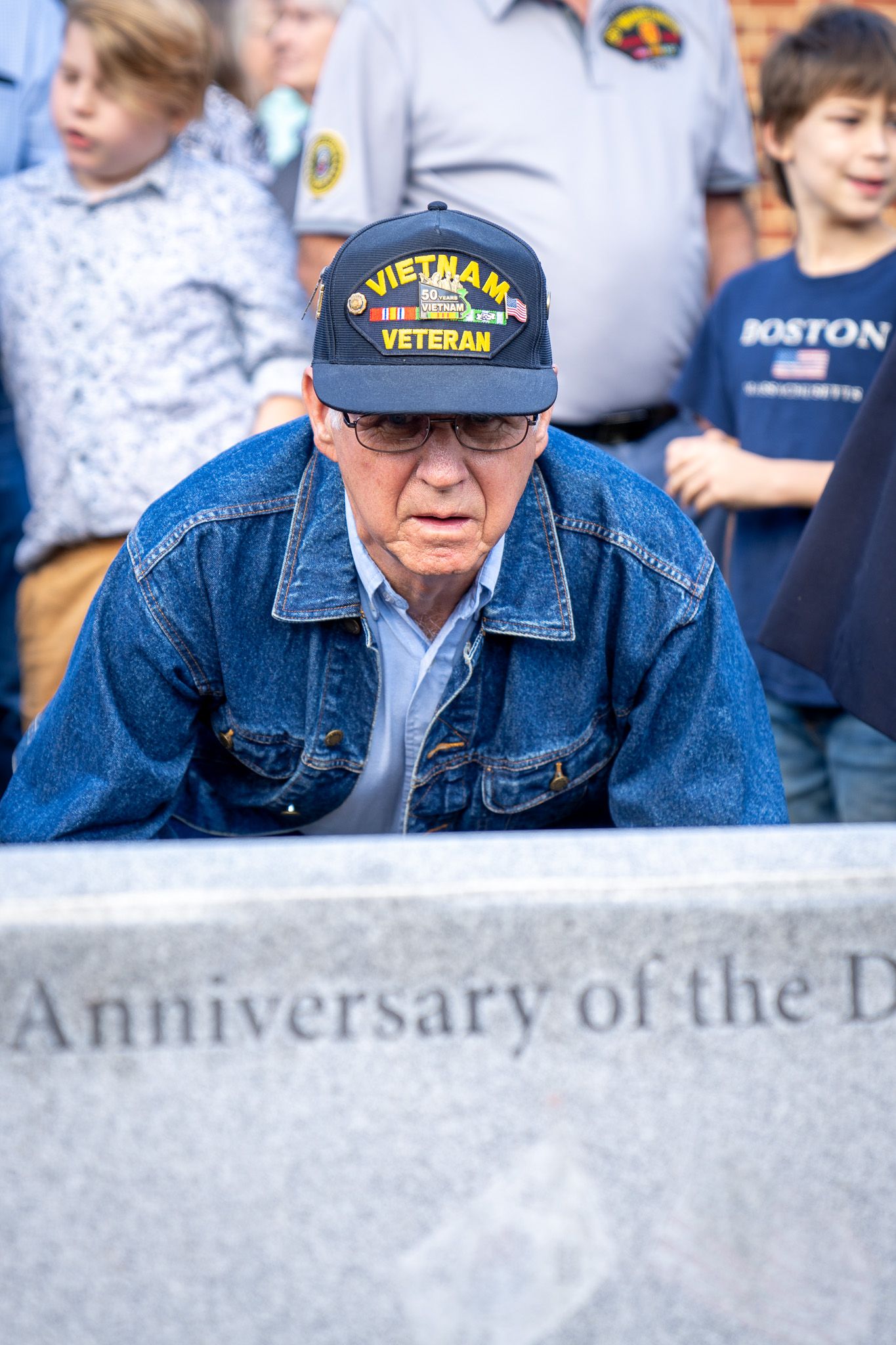 A Vietnam veteran wearing a navy blue military cap and denim jacket looks down at a memorial stone.
