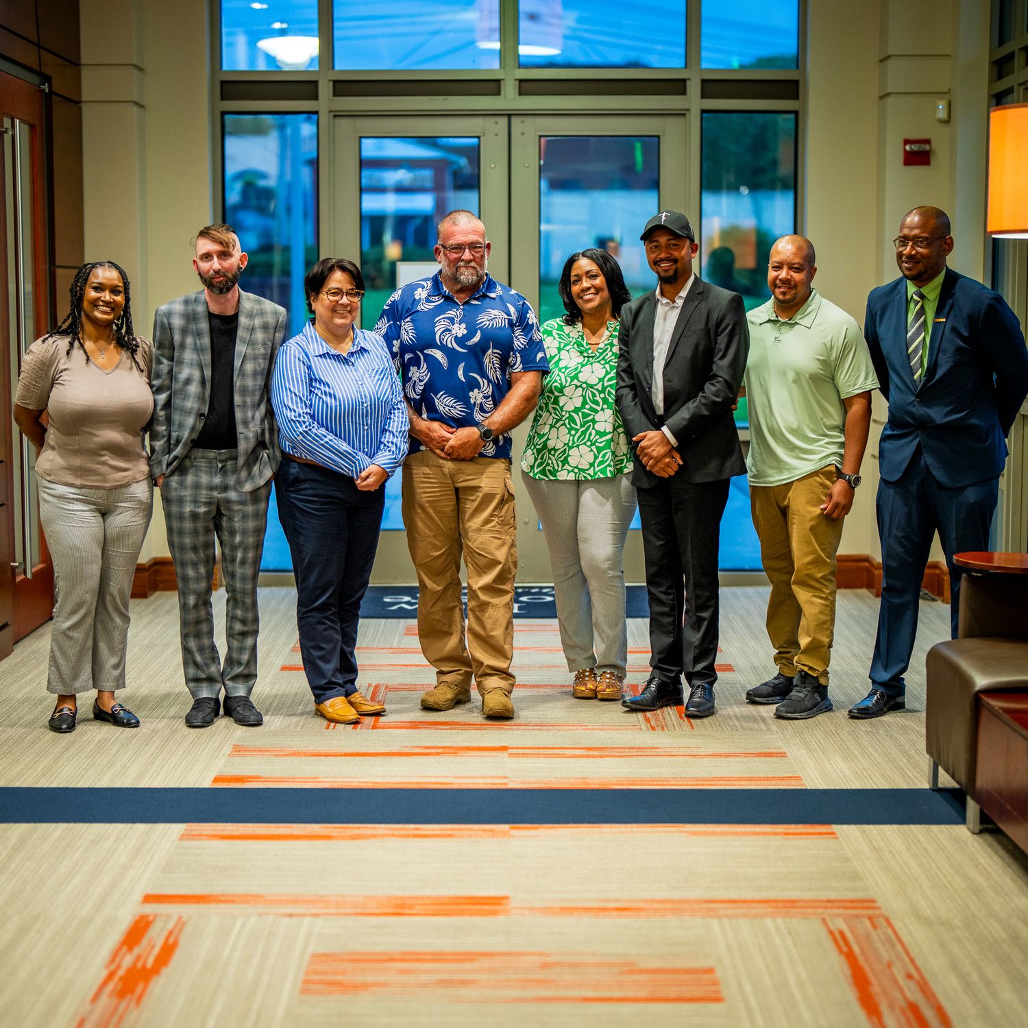 Professional group photo of eight diverse colleagues standing together in a modern office building lobby.