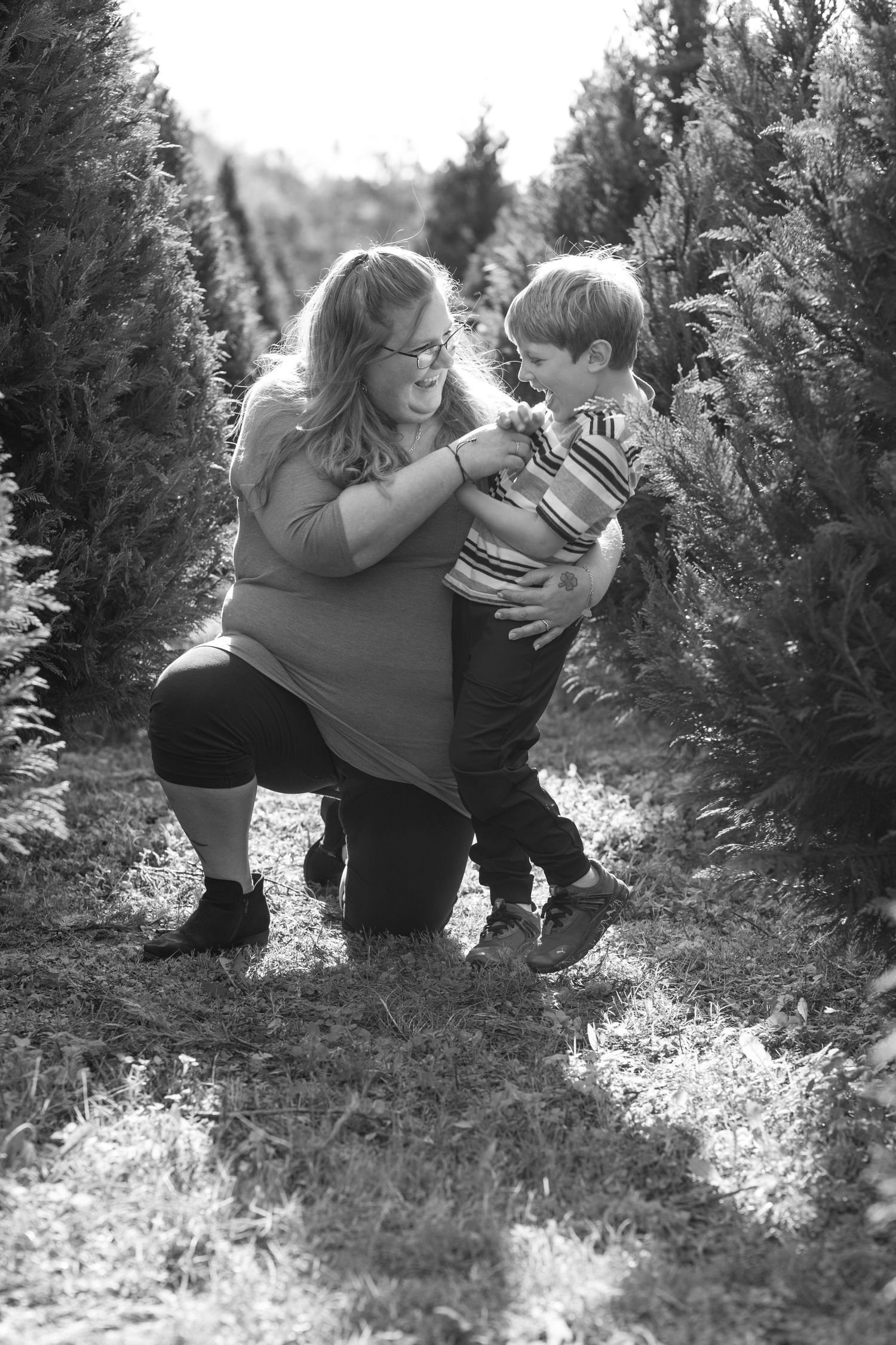 Black-and-white portrait of woman crouching down between Christmas trees, smiling warmly — holiday photography in York County, SC.