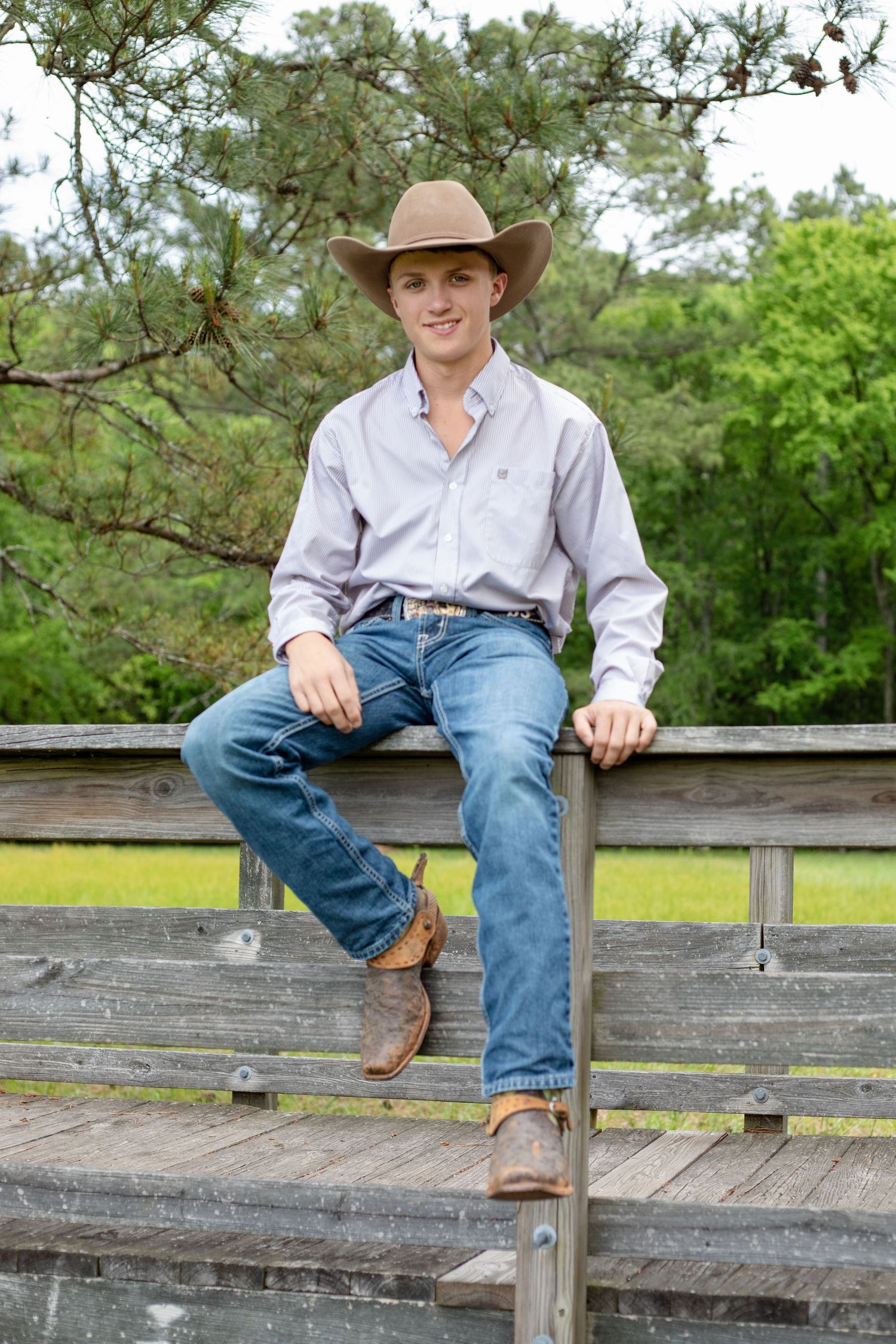 High school senior in cowboy hat and jeans sitting on fence for rustic outdoor graduation portraits in York County, SC.