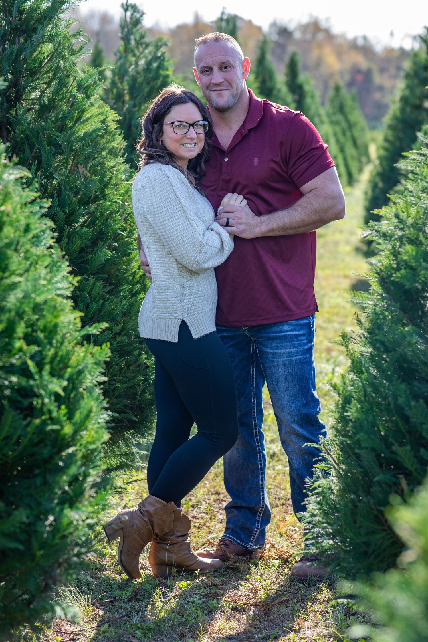 Couple embracing between rows of Christmas trees, wearing cozy winter outfits — romantic holiday portraits in McConnells, SC.