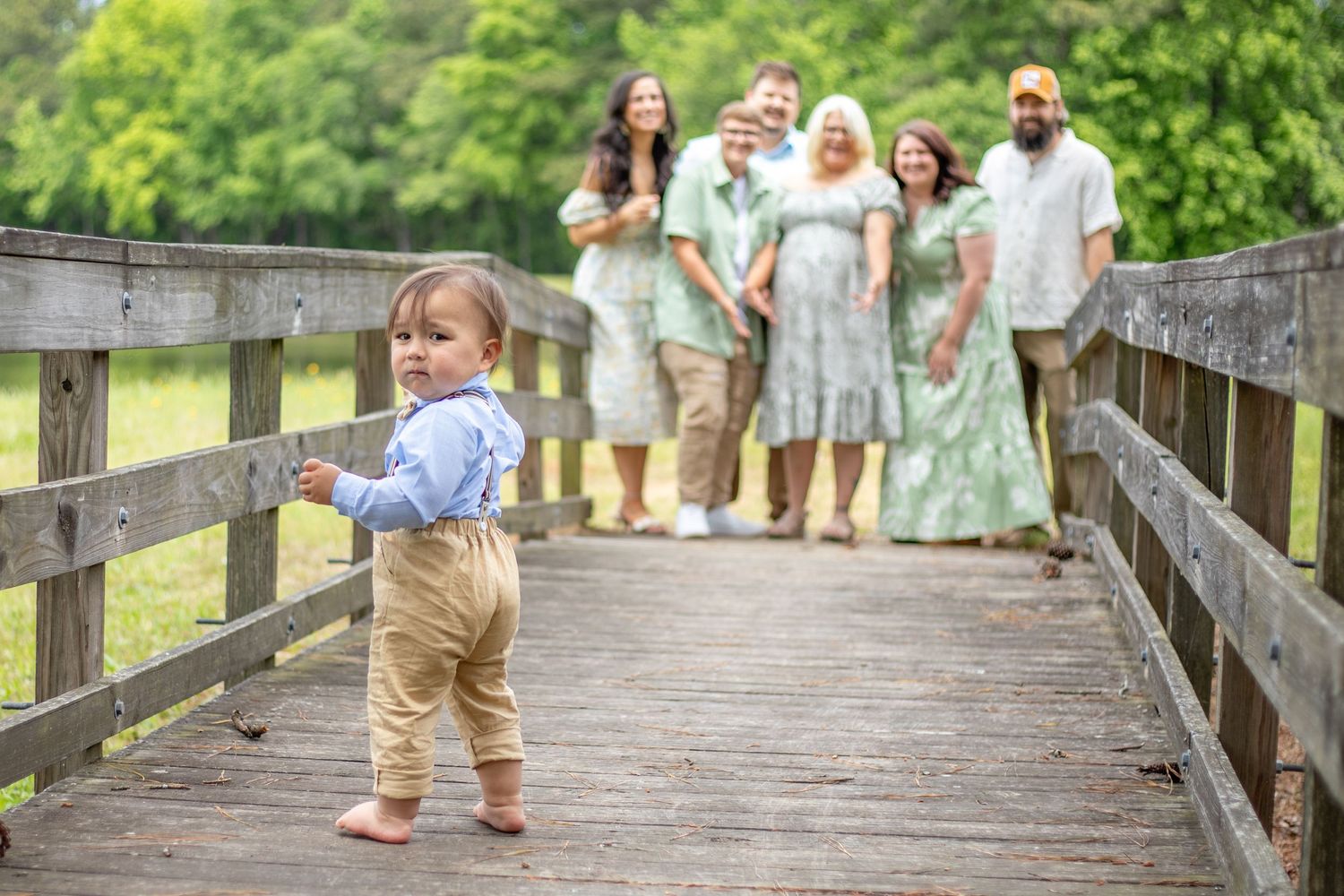 Toddler walking on wooden bridge with family in soft pastel outfits in the background — outdoor family photography in McConnells, SC