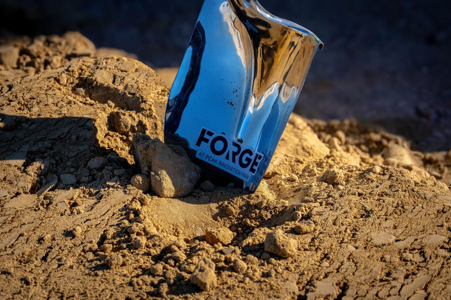 A blue Forge-branded shovel blade pressed into sandy soil during a groundbreaking ceremony.