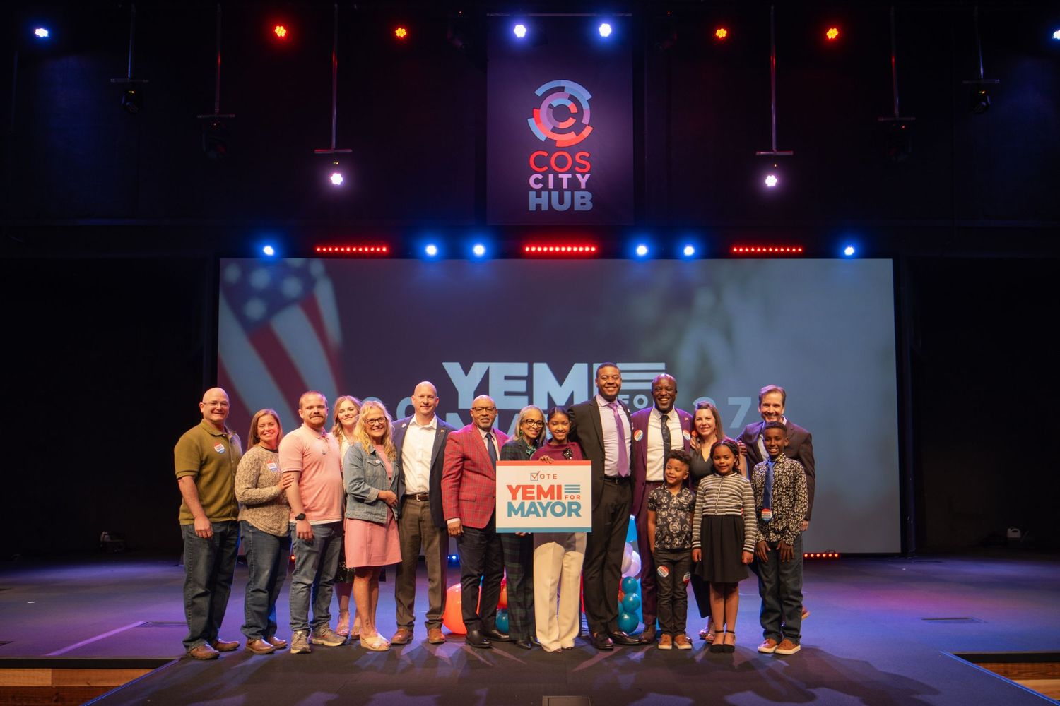 Group of people posing on stage at COS City Hub with a YEM for Mayor campaign banner.