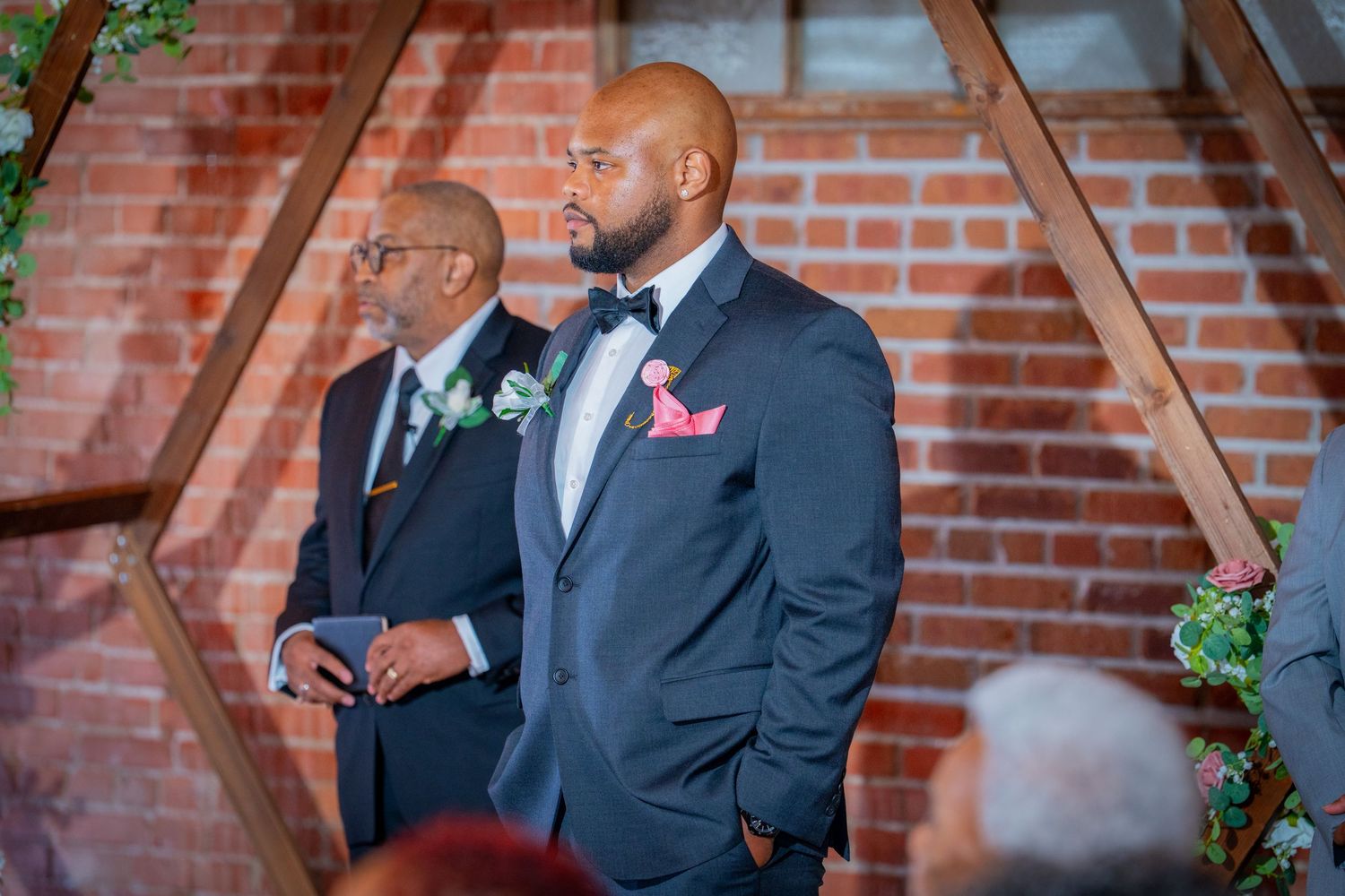 Groom in navy suit with pink boutonniere stands at wedding altar beside older man in brick venue.