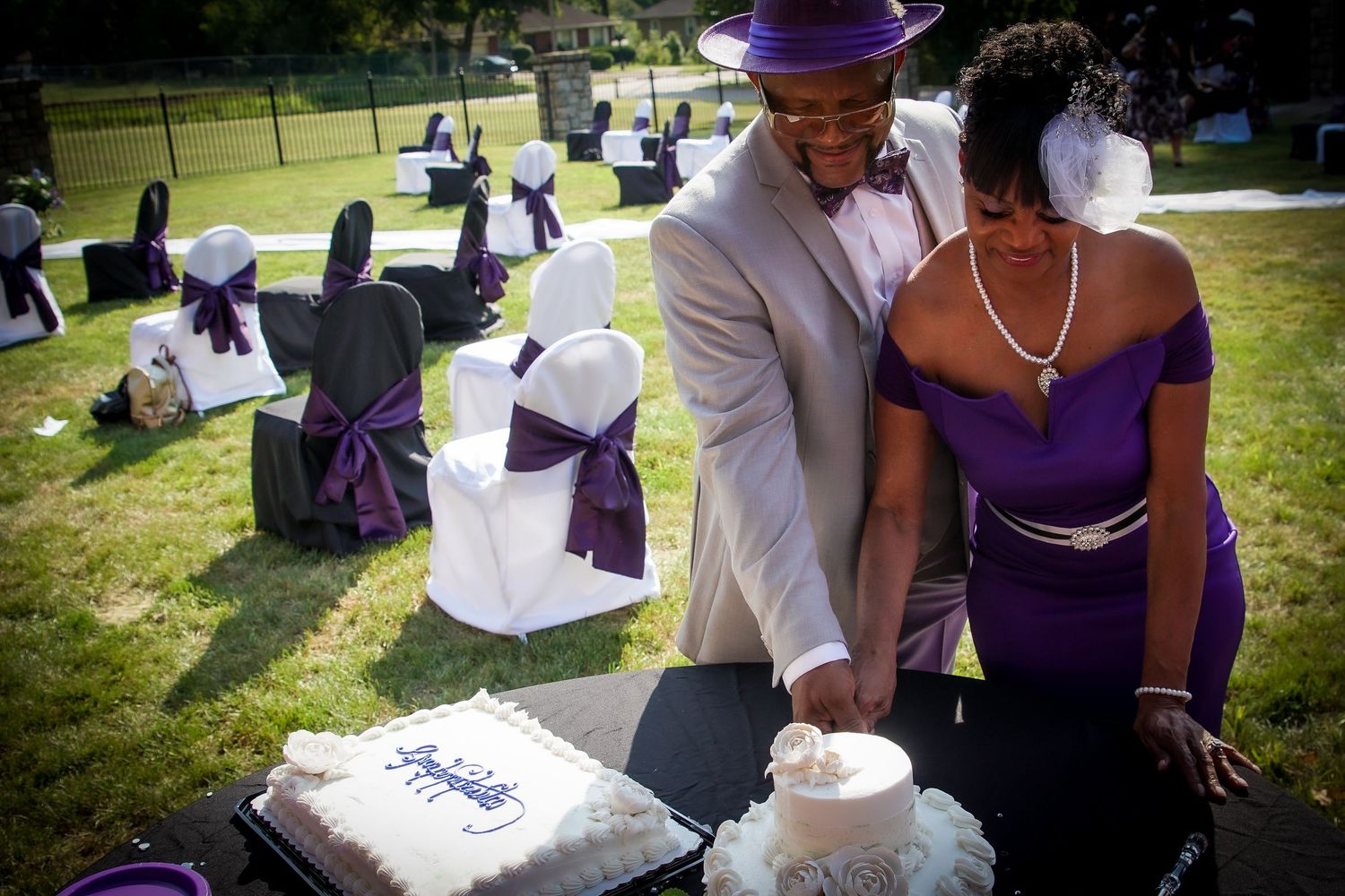 A couple cuts their wedding cake at an outdoor reception with purple and white decorations.
