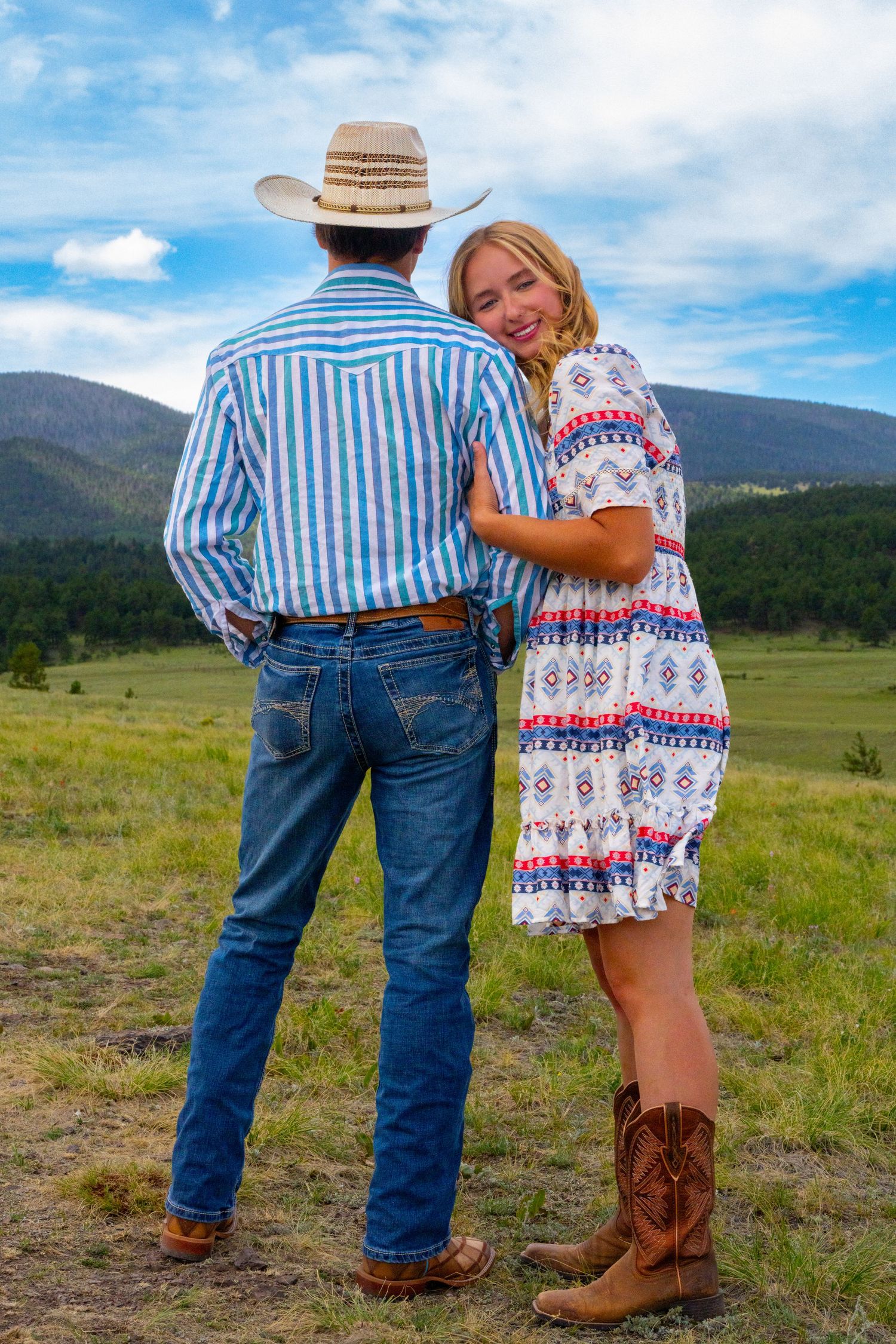 A couple in western attire stands together on a hillside overlooking mountains and meadows on a sunny day.