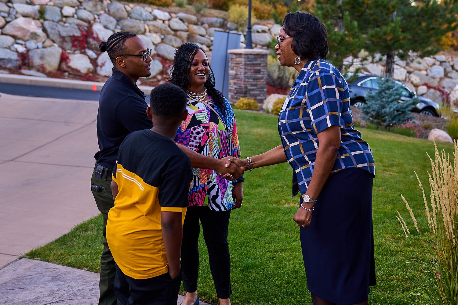A group stands together having an animated conversation outdoors on a landscaped path during evening hours.