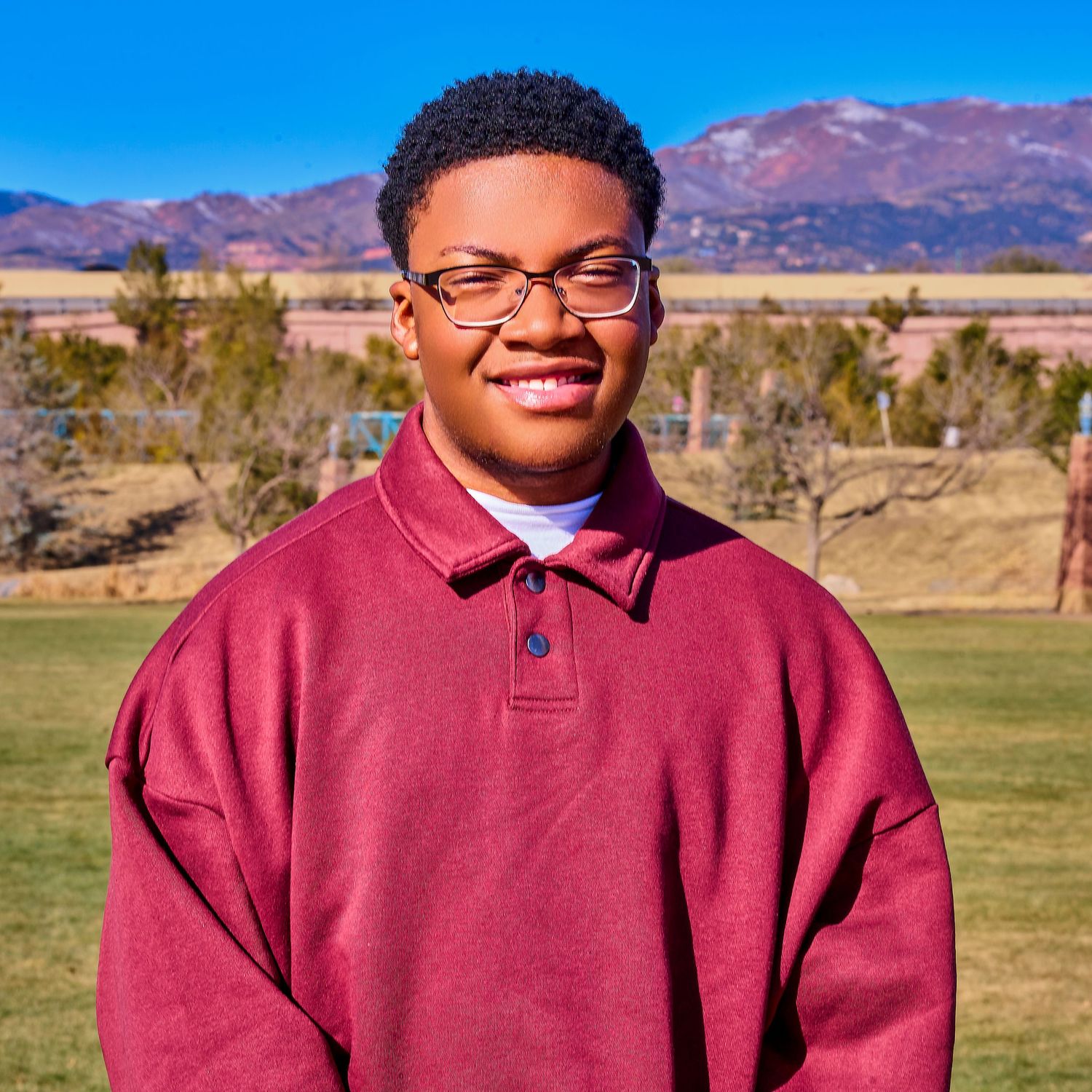 Young person in a burgundy polo shirt smiling outdoors with mountains in the background.