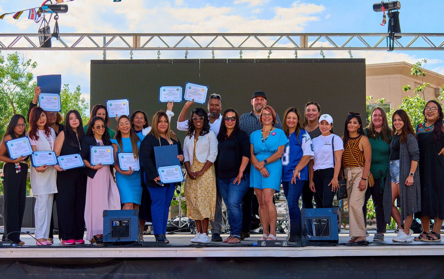 Large group of diverse professionals posing together on outdoor stage with certificates.