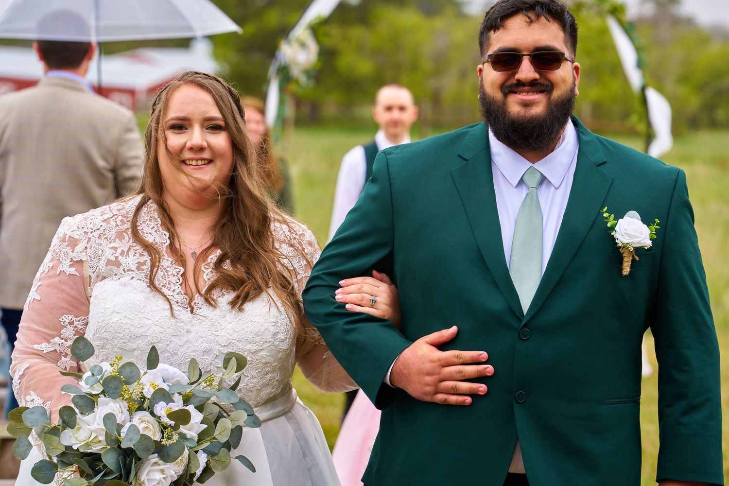 A couple looks joyful walking together at an outdoor wedding ceremony with the bride holding a white floral bouquet.