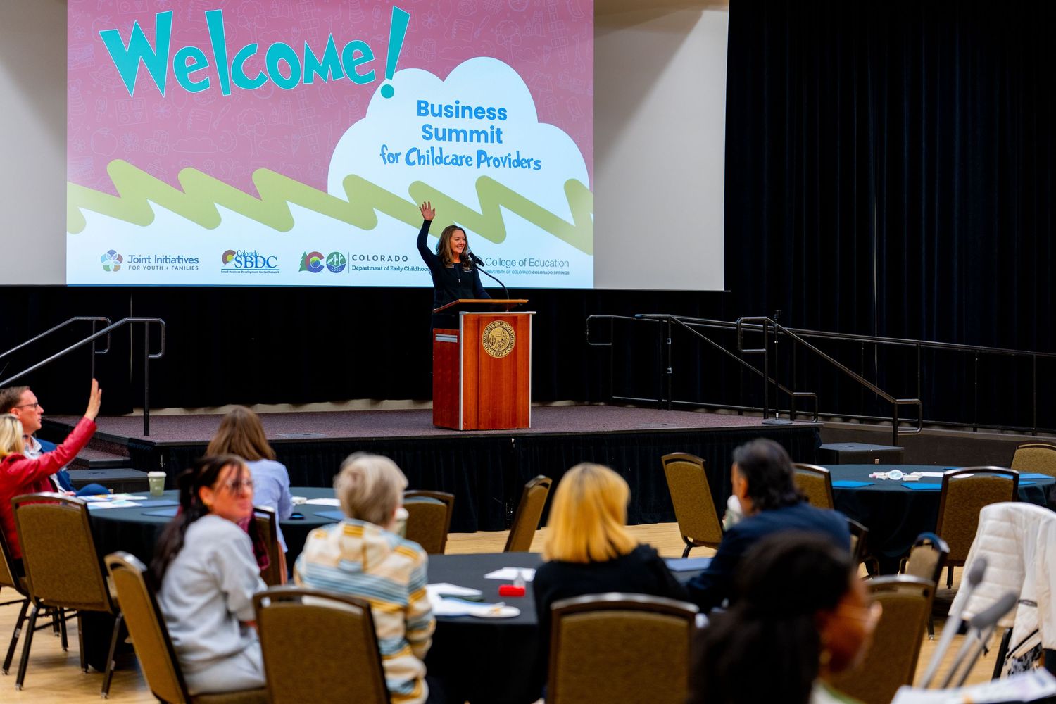 Speaker presents at Business Summit for Childcare Providers welcome session in a conference hall with seated attendees.