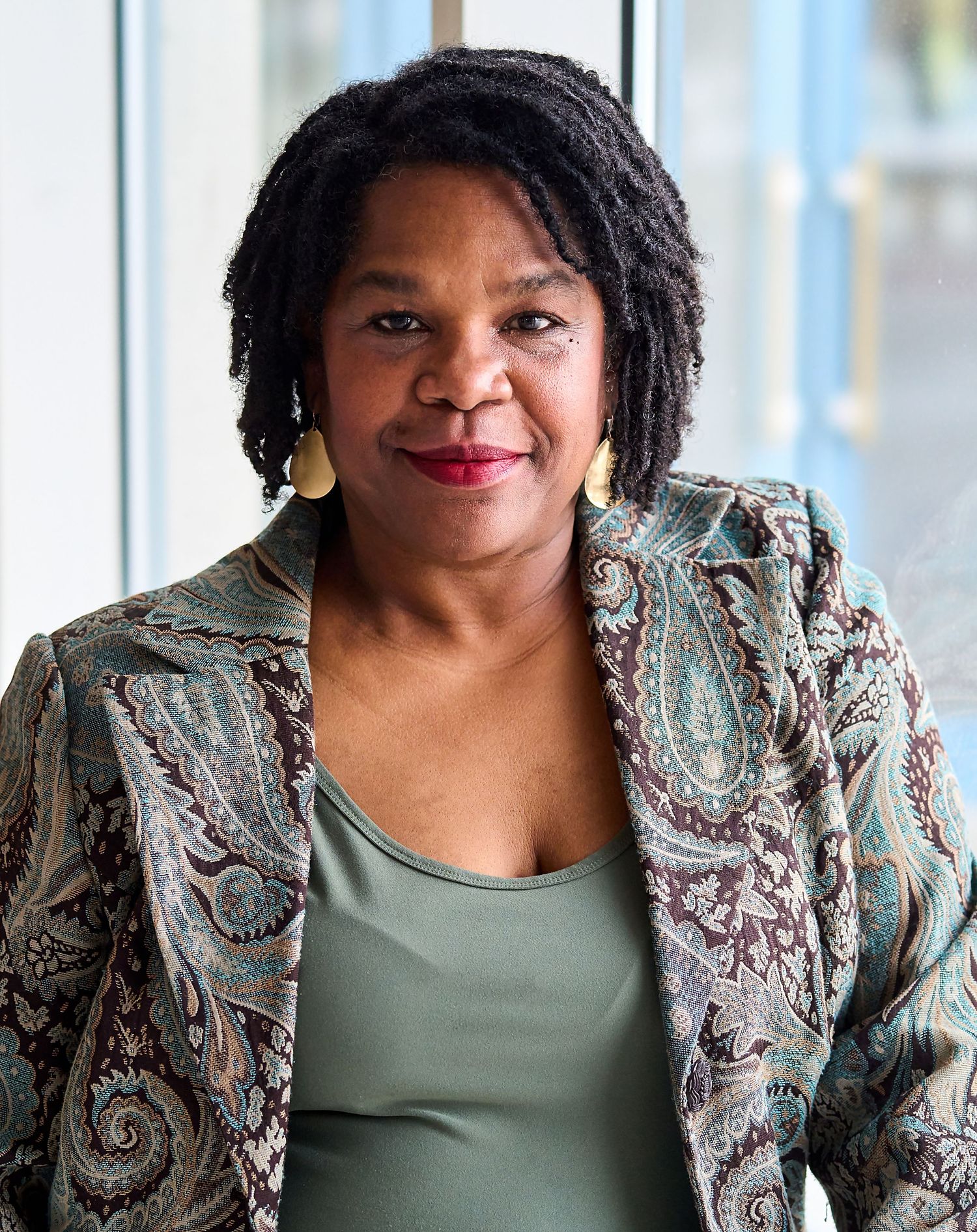 Woman wearing patterned blazer and green top stands in front of window with natural lighting.