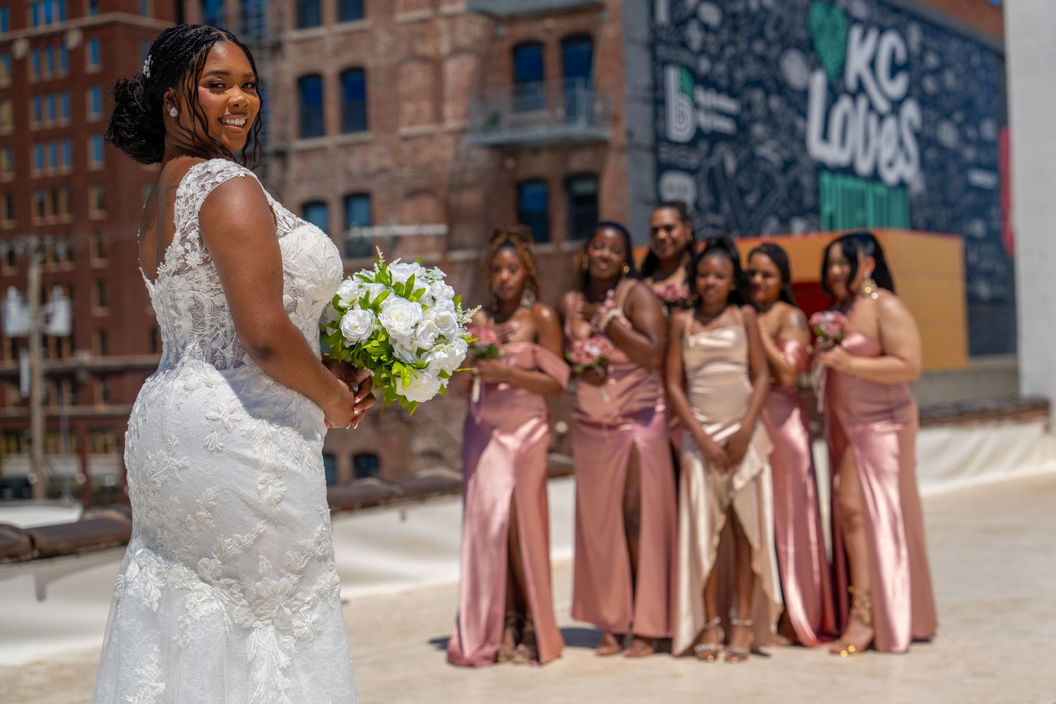 Bride in lace gown holds white bouquet with bridesmaids in pink dresses posing before KC Loves mural.