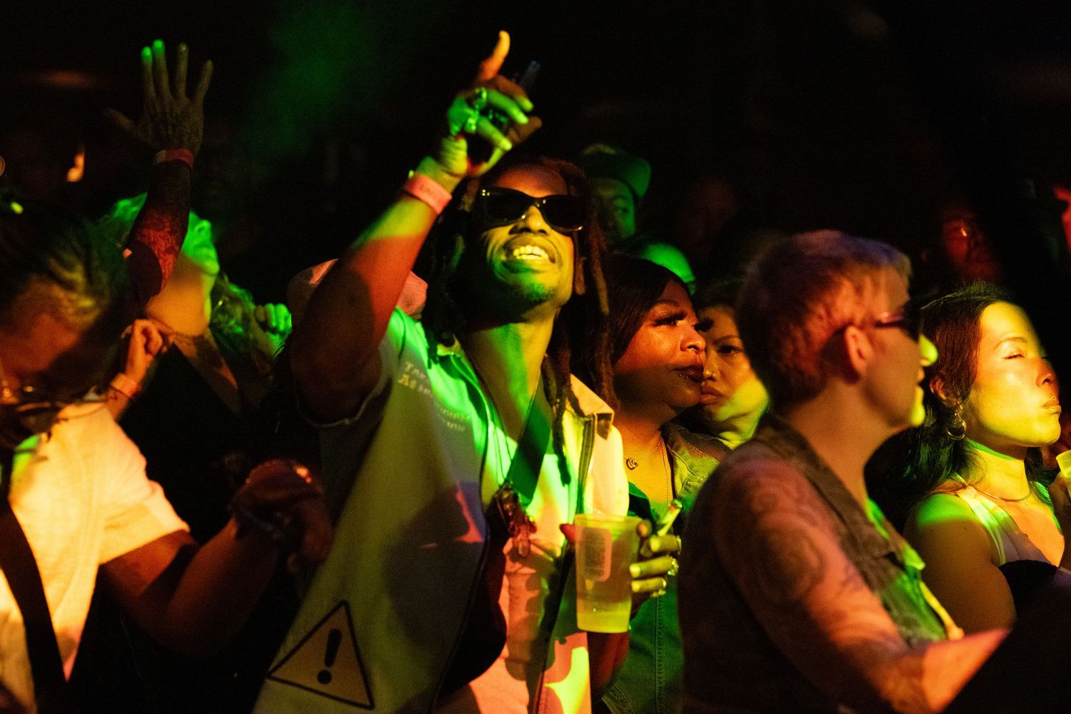 Concert crowd watches performers on stage under colorful green and yellow lighting at a nighttime music event.