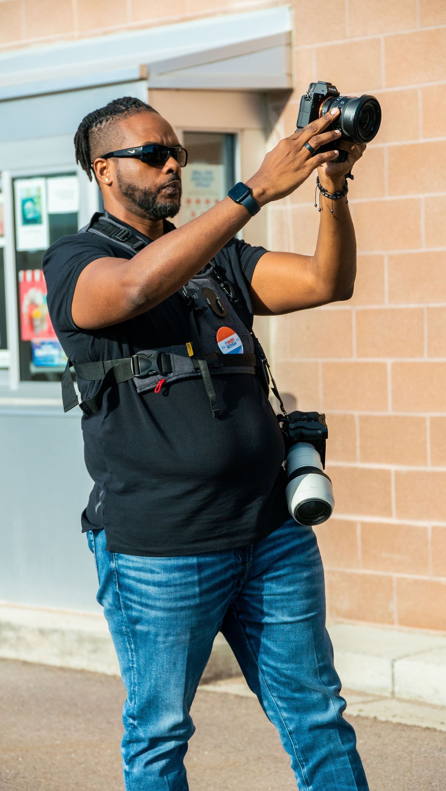 Professional photographer with sunglasses and tactical vest holding up a camera against a beige wall.
