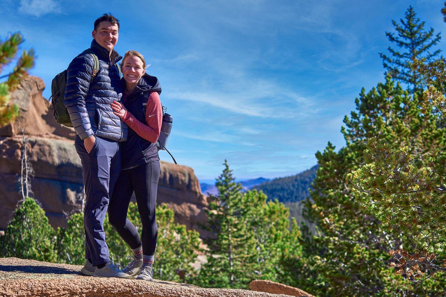 Couple standing together on rocky overlook with mountain vista in background.