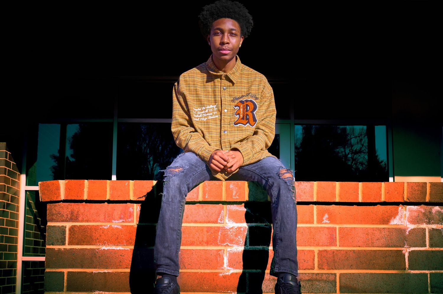 Young man in yellow plaid jacket and ripped jeans sitting on a brick wall outdoors.