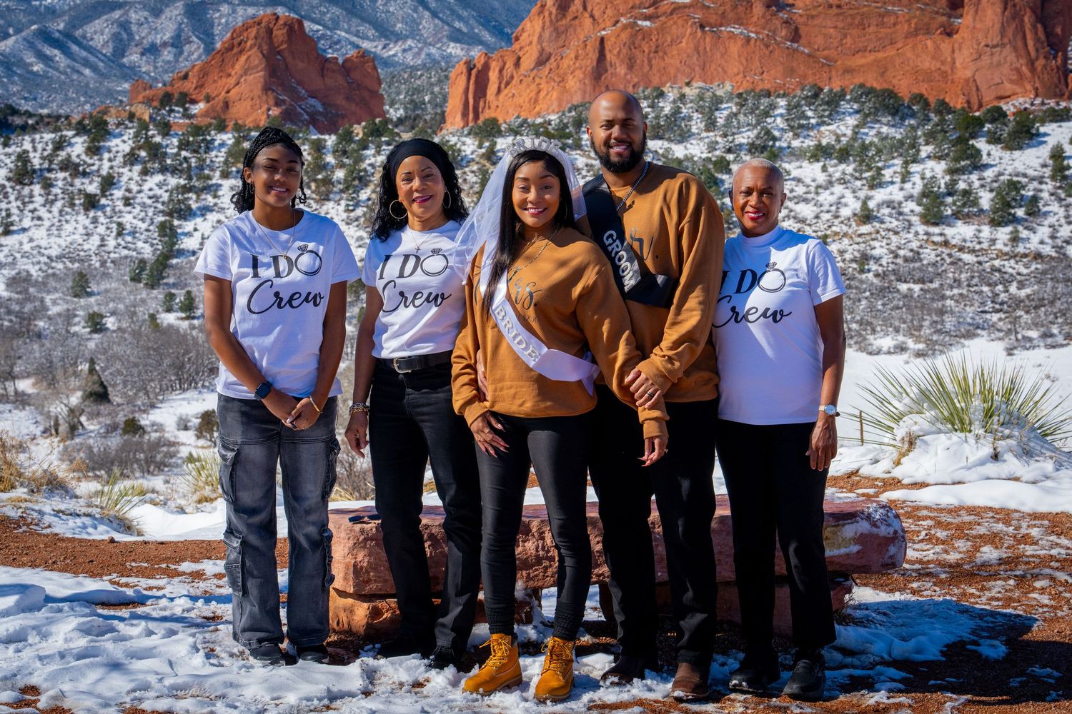 Group of five people in I Do Crew shirts posing at Garden of the Gods, Colorado, with snowy red rock formations.