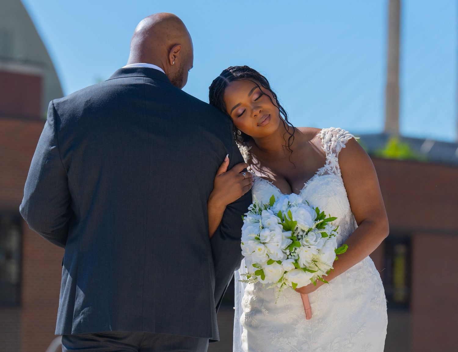 Bride in white lace gown holding white bouquet shares intimate moment with groom in navy suit outdoors.