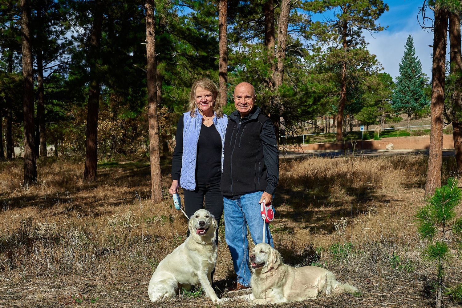 Couple poses with their two white dogs in a forest setting on a sunny day.