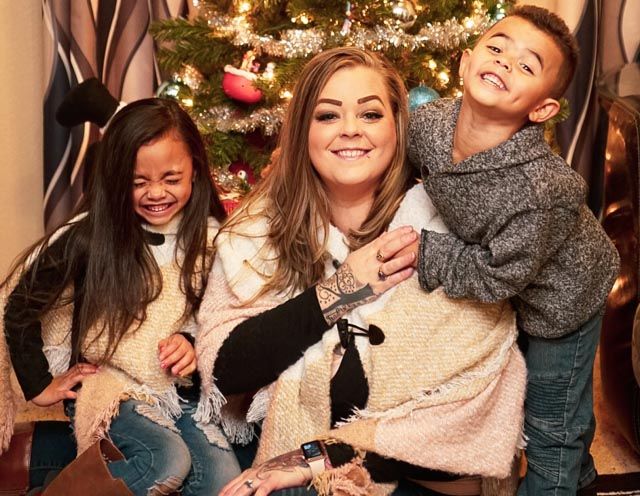 Three people laughing together in front of a decorated Christmas tree with twinkling lights.