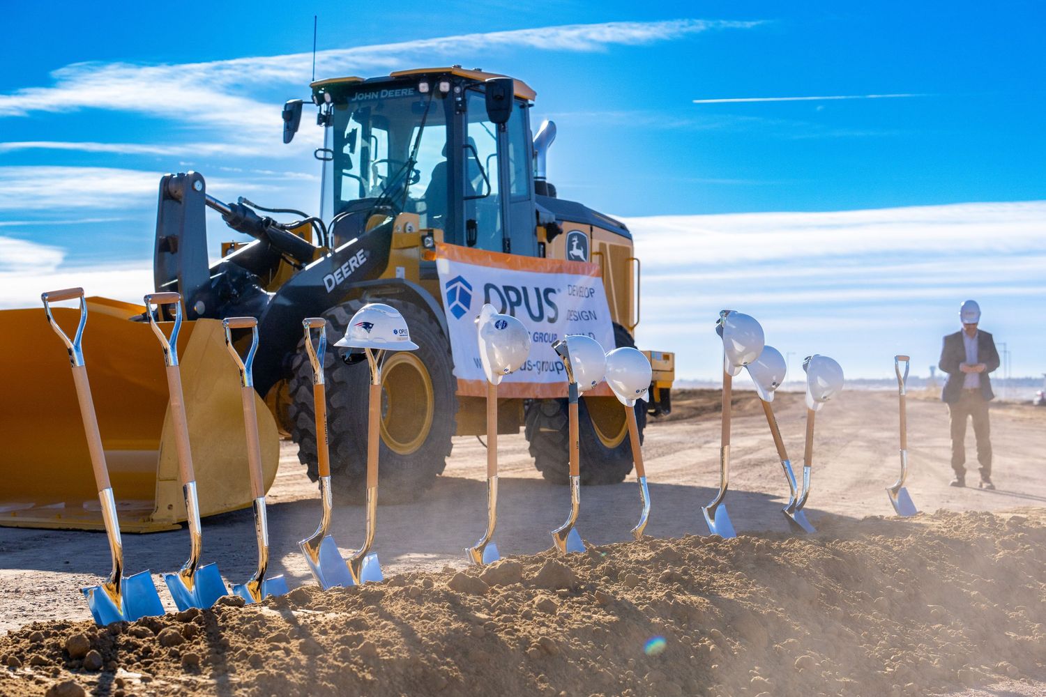 Opus groundbreaking ceremony with yellow bulldozer and golden shovels in dirt under blue sky.