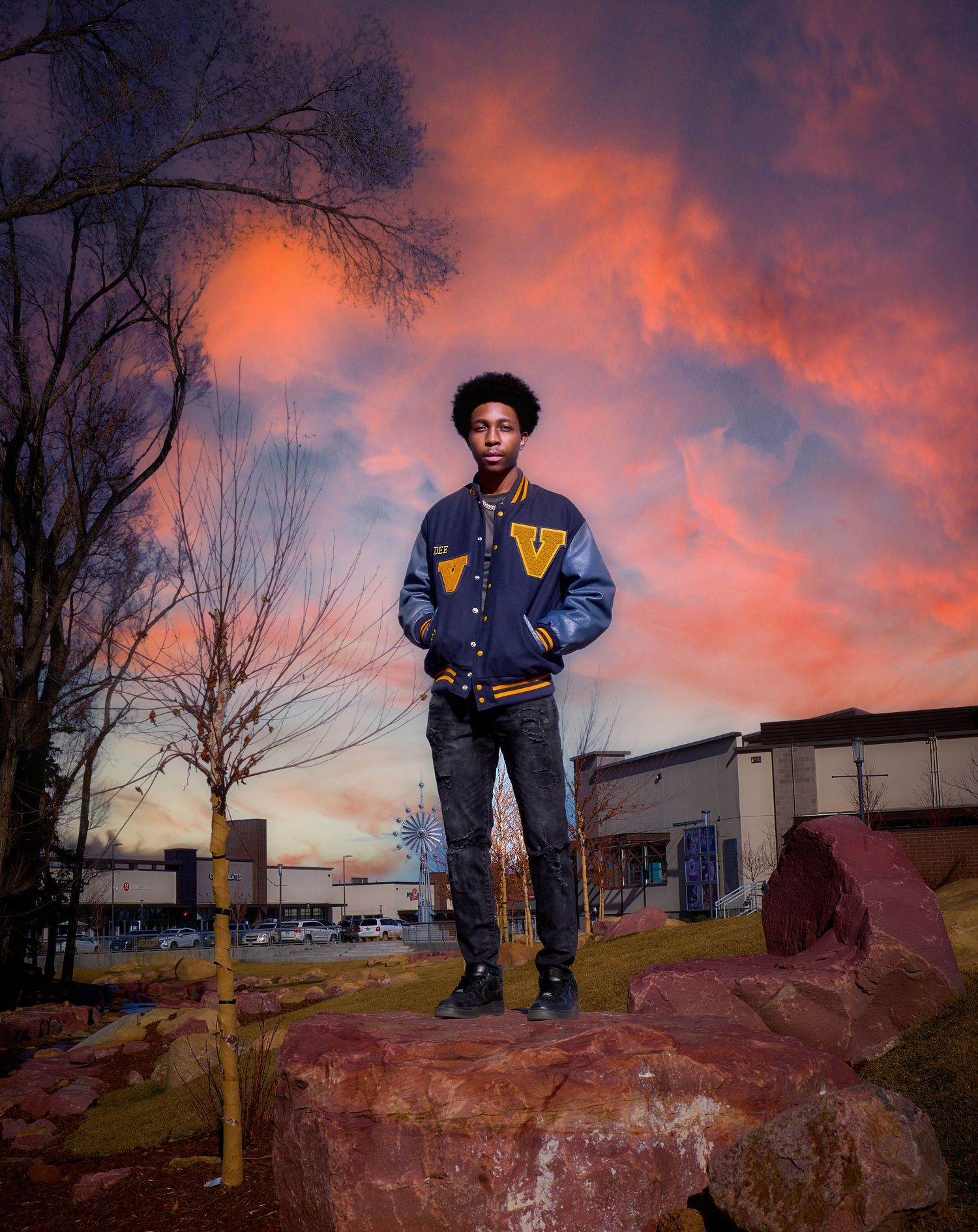 A person stands confidently on rocky terrain during sunset while wearing a varsity jacket and jeans.