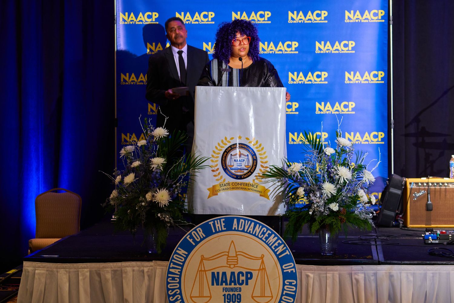 Speakers at NAACP podium with organization logos and floral arrangements on stage.