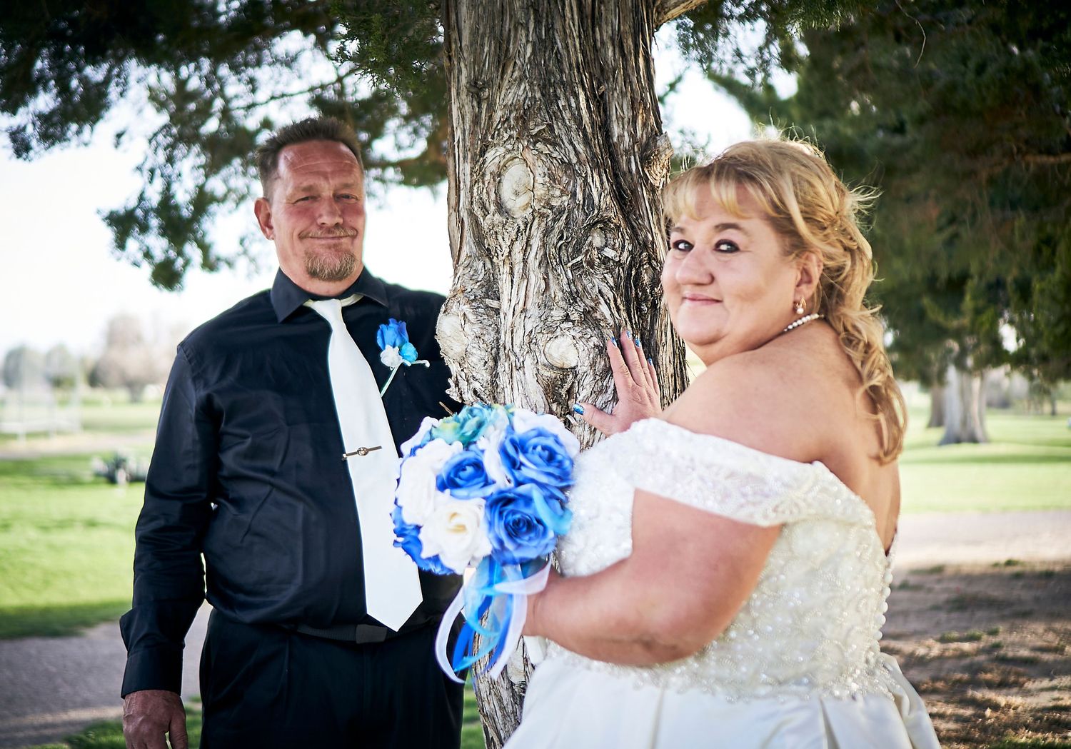 Wedding photo of couple by tree with bride holding blue bouquet.