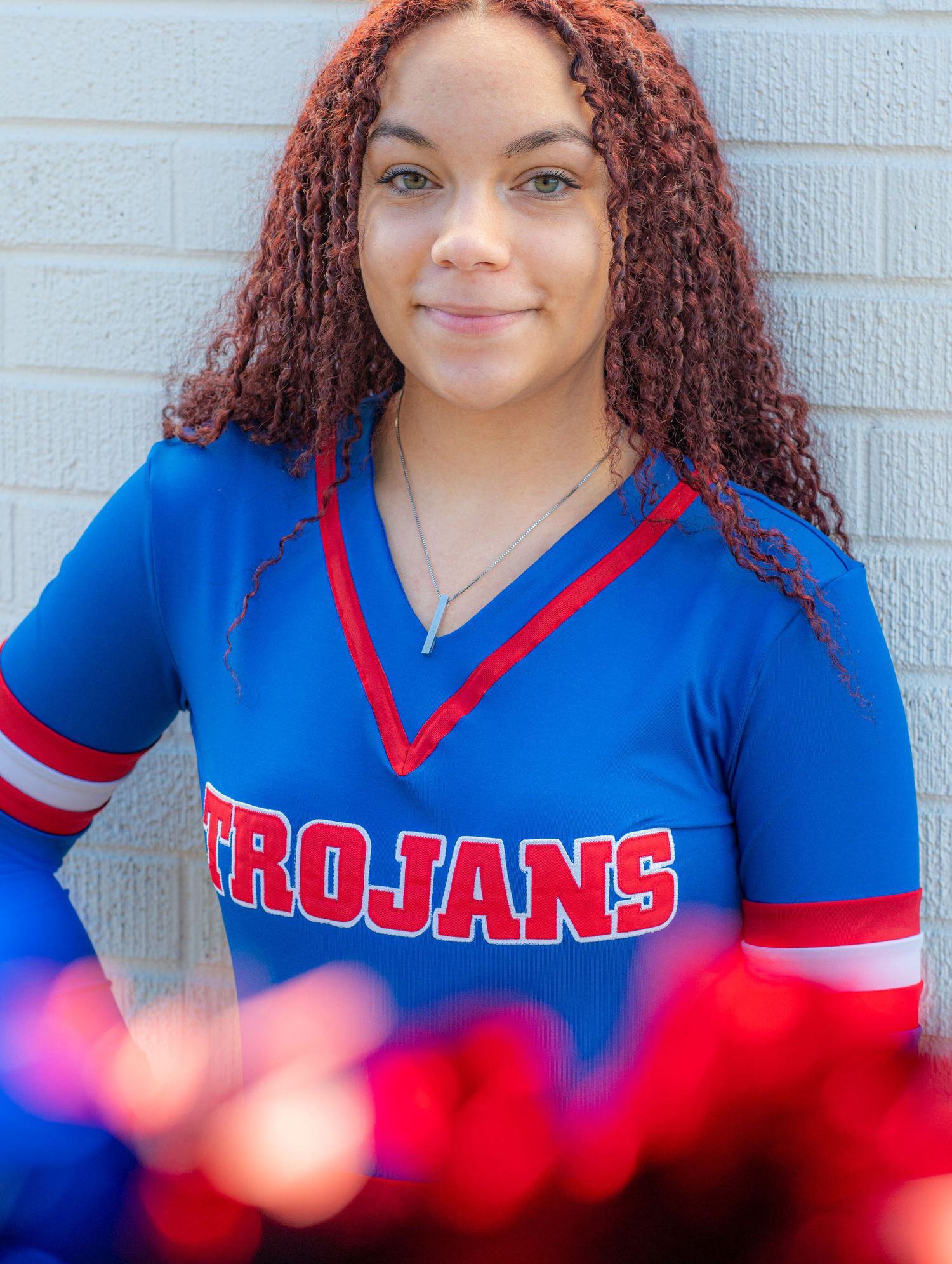 Student wearing blue and red Trojans cheerleading uniform with curly hair against white wall.
