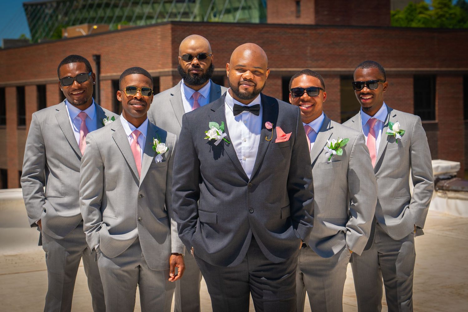 Groom in dark gray suit with bow tie poses with groomsmen in light gray suits and pink ties outdoors.