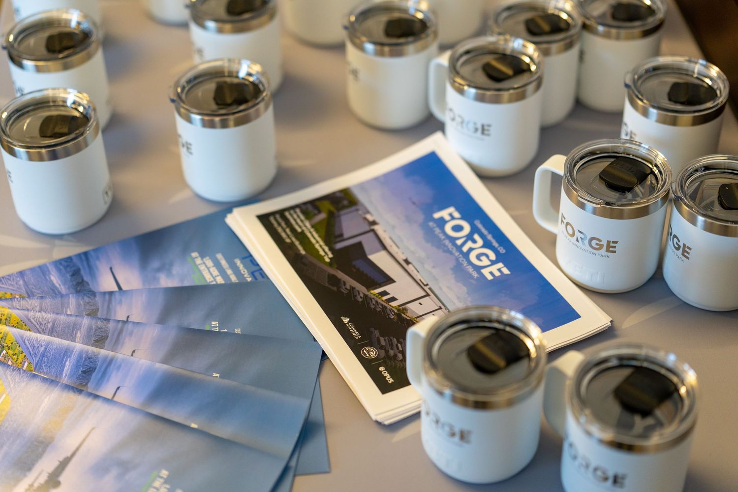 White branded Forge mugs and brochures displayed on a table at a corporate event or conference.