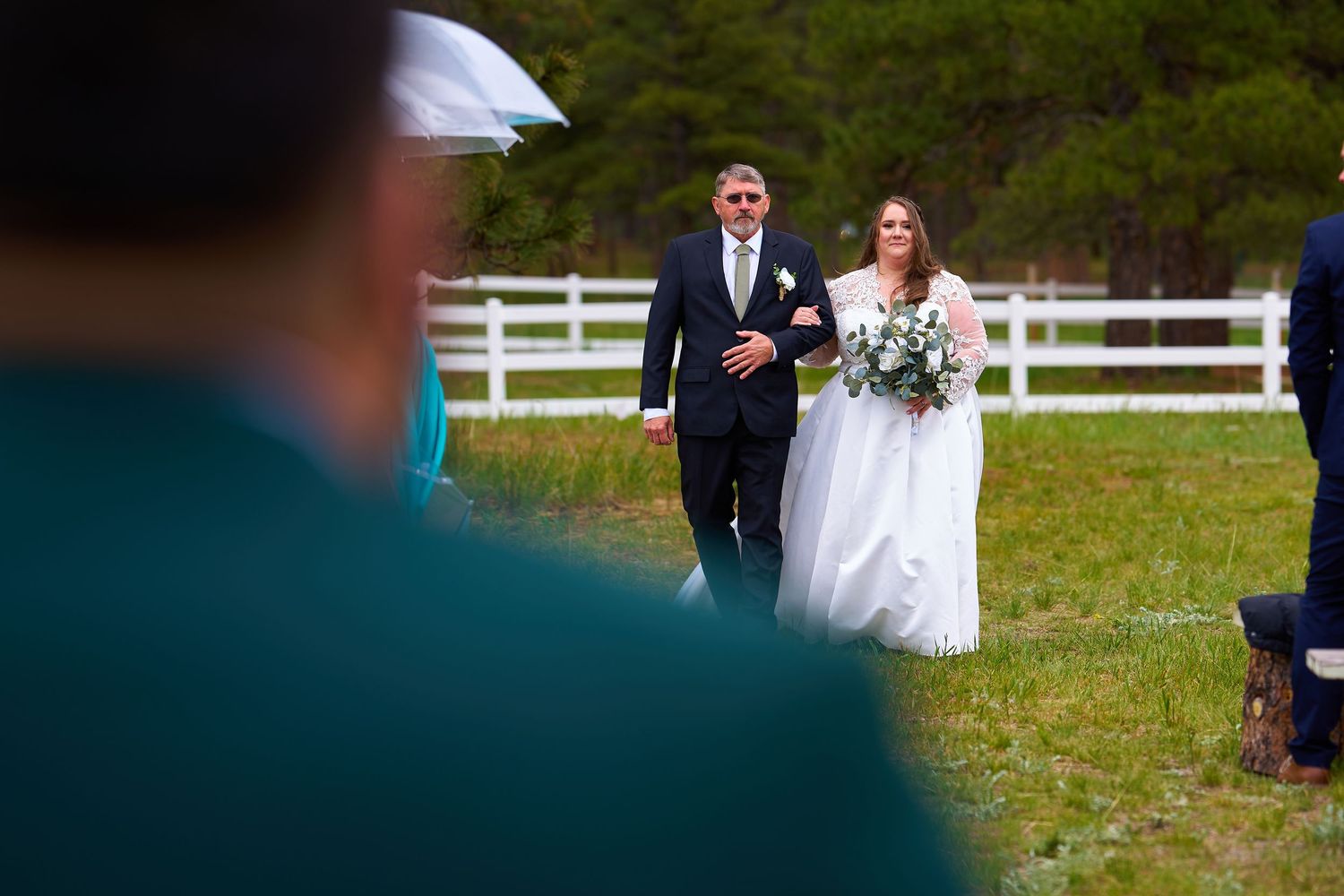 Bride in white dress walks down grassy aisle with escort past white fence on wedding day.