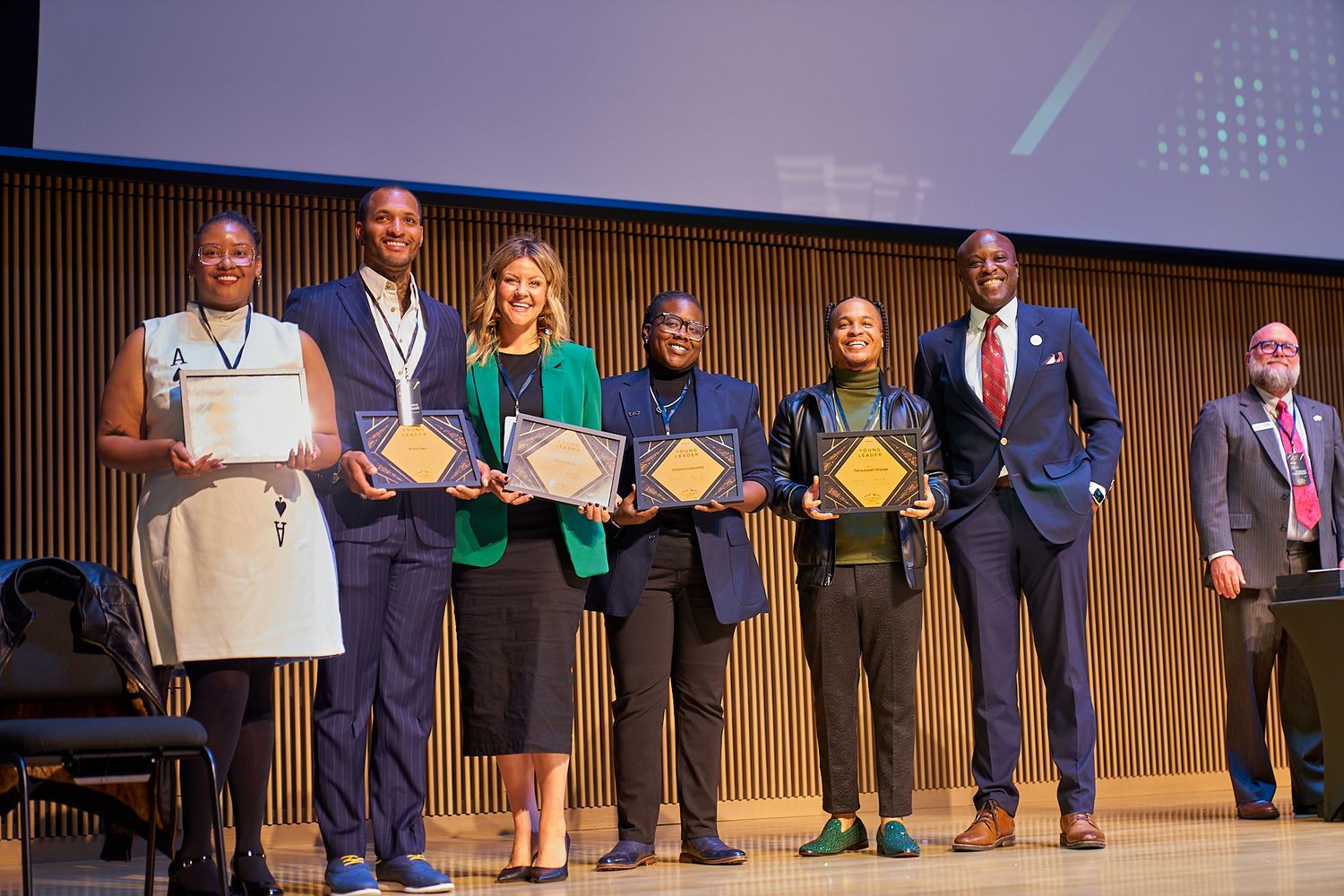 A group of award recipients stand on stage holding certificates and plaques during a formal ceremony.