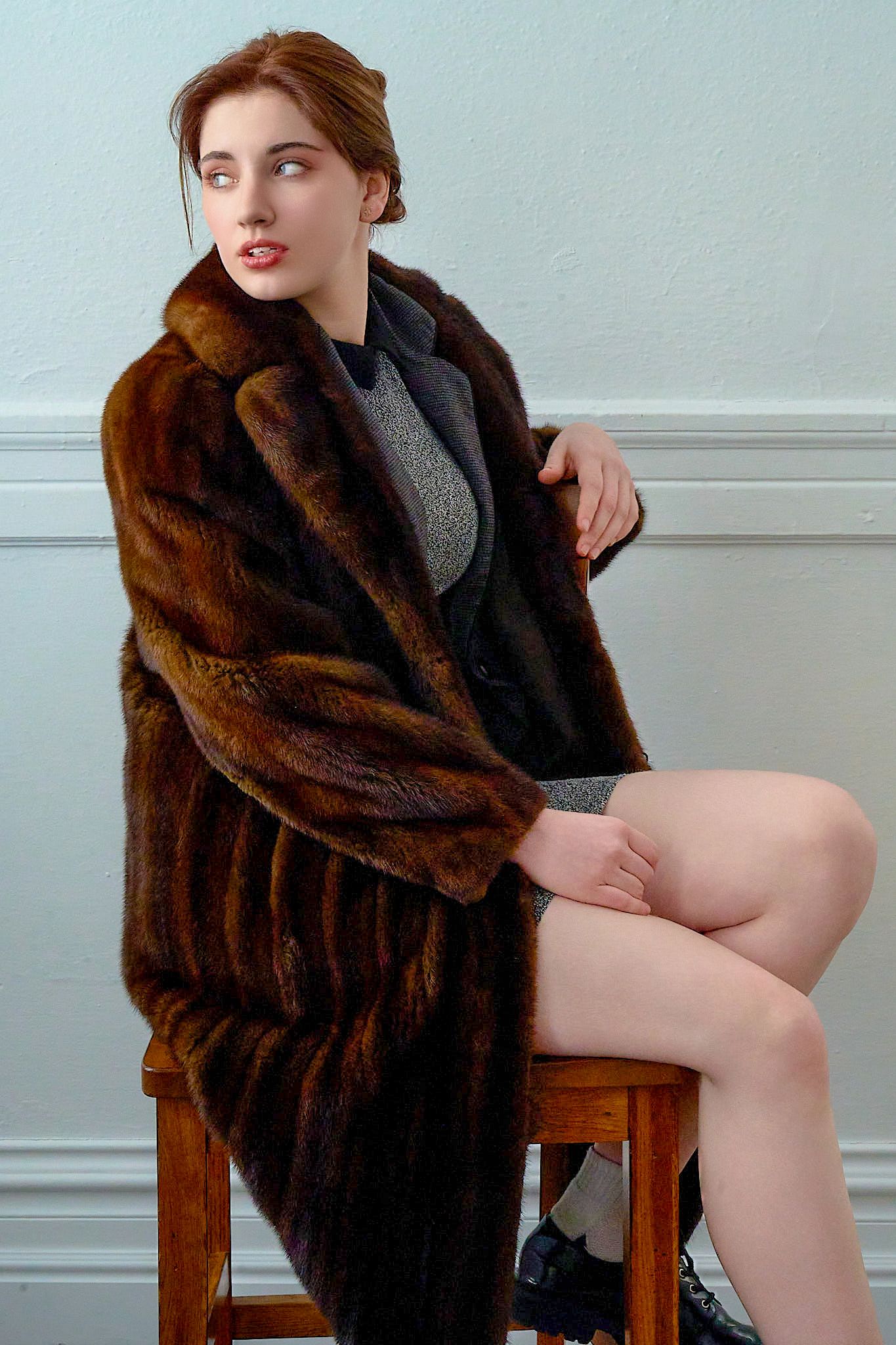 Fashion model wearing brown fur coat sitting on wooden stool against light wall.