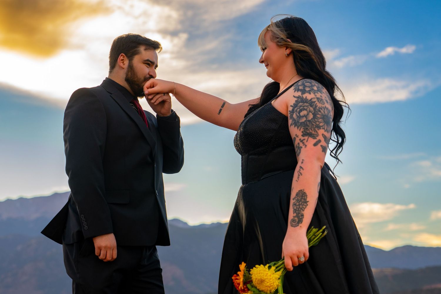 A couple embraces during a sunset photoshoot in front of mountain scenery with dramatic clouds.