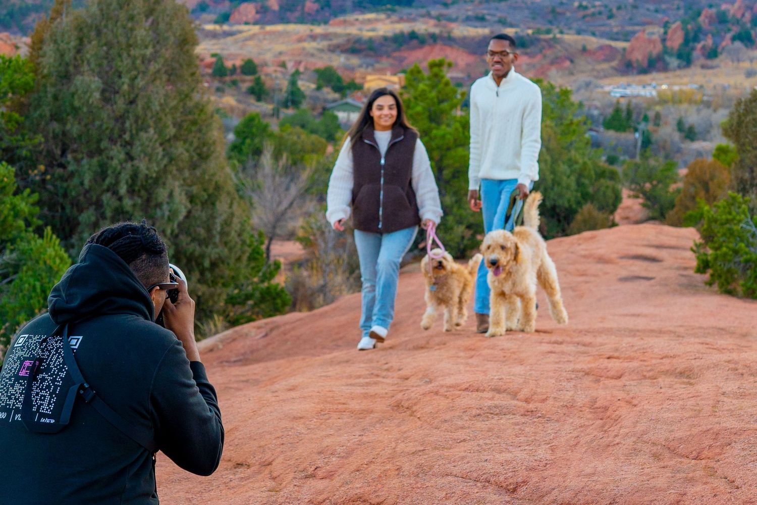 A photographer captures people walking their dogs on a red rock trail with scenic mountain views in the background.