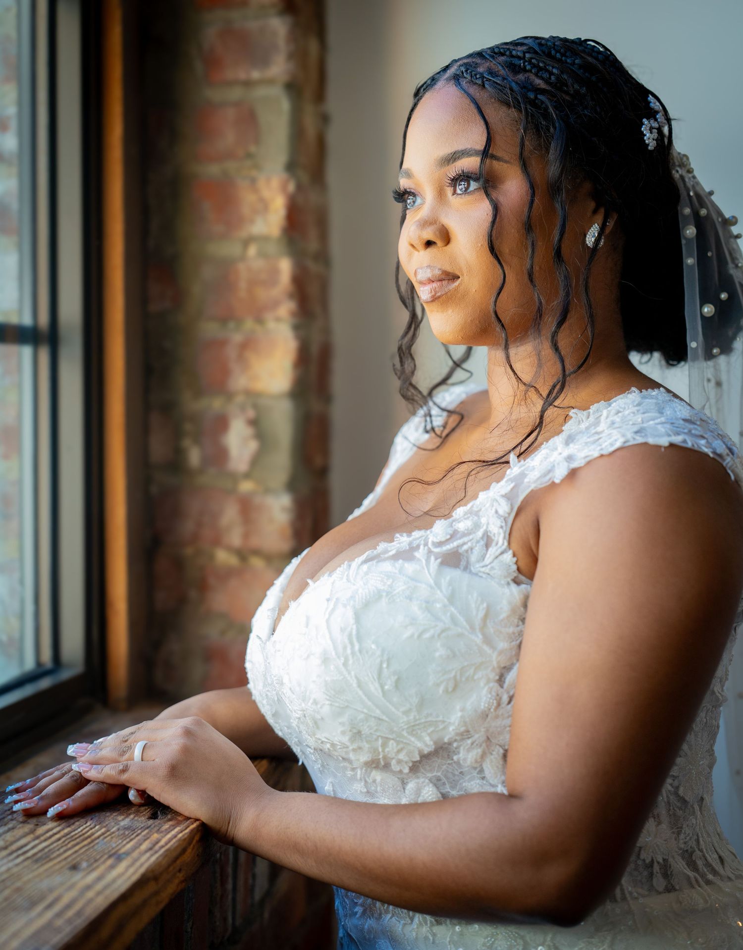 A Black bride in a white lace gown gazes thoughtfully out a window, wearing a veil and elegant makeup.
