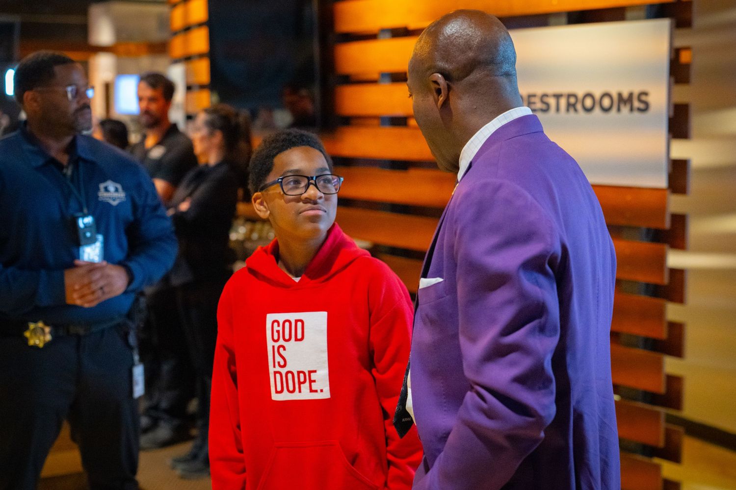 A youth in a red God Is Dope hoodie converses with a man in a purple suit near a restrooms sign.