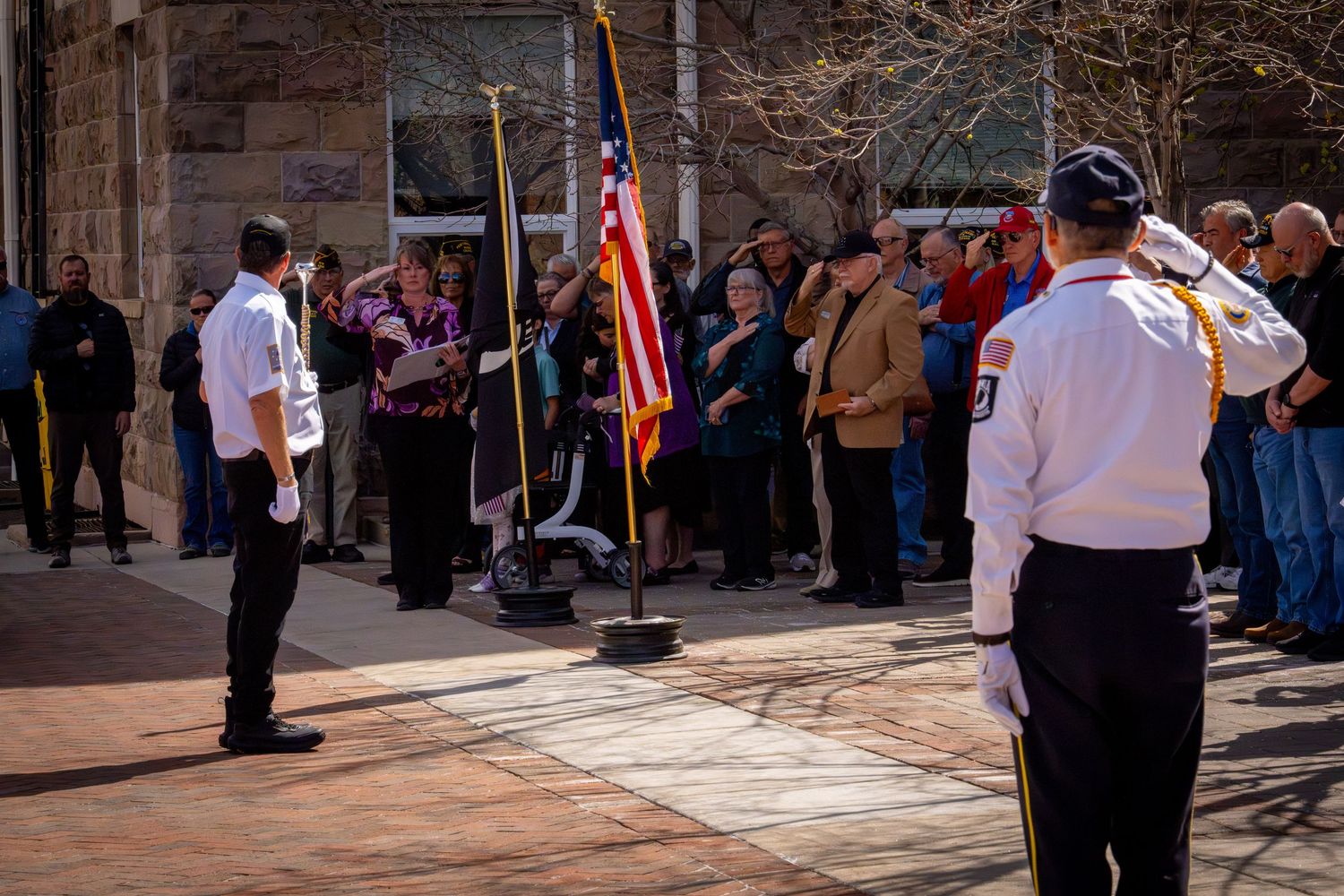 Veterans salute the American flag during an outdoor military ceremony with a crowd of attendees.