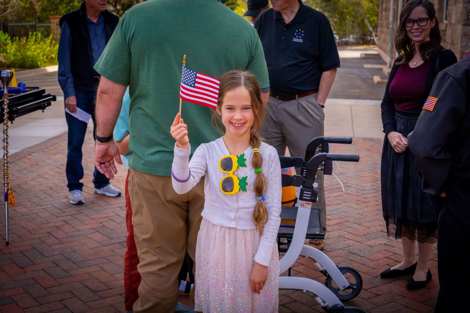 Young girl smiling and waving an American flag at an outdoor patriotic ceremony.